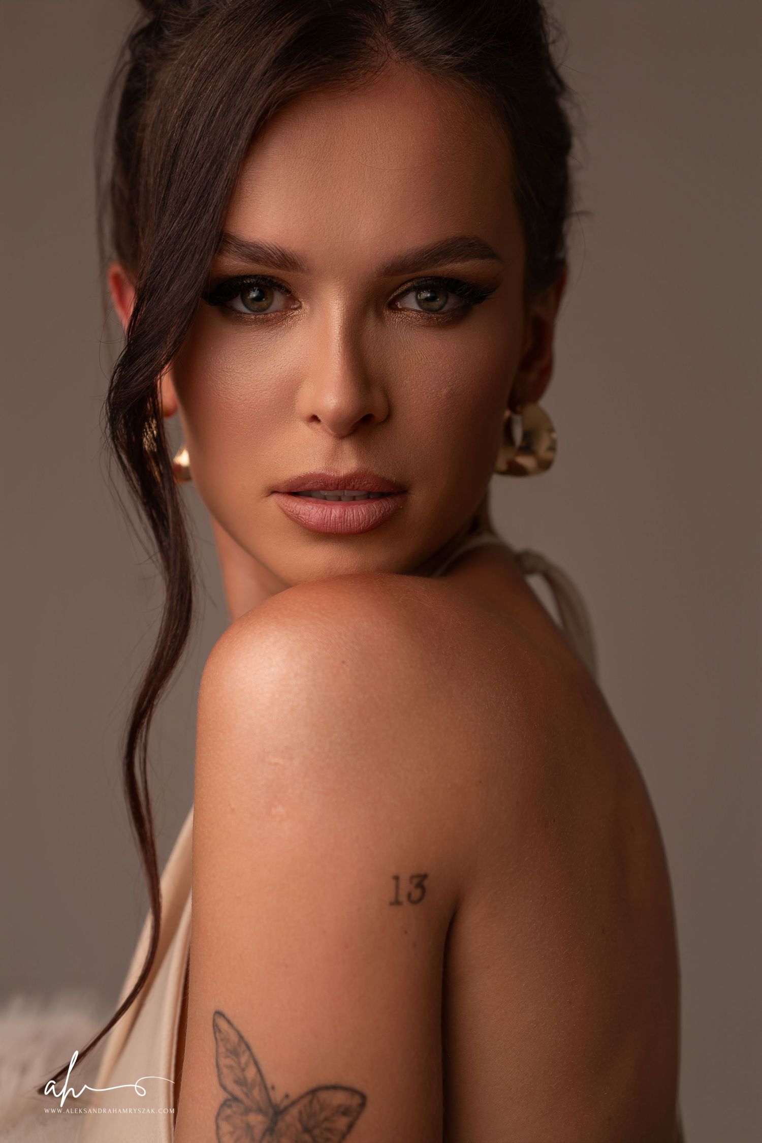 Beautiful portrait with dramatic lighting showcasing bare shoulders, butterfly tattoo, and gold hoop earrings against neutral backdrop.