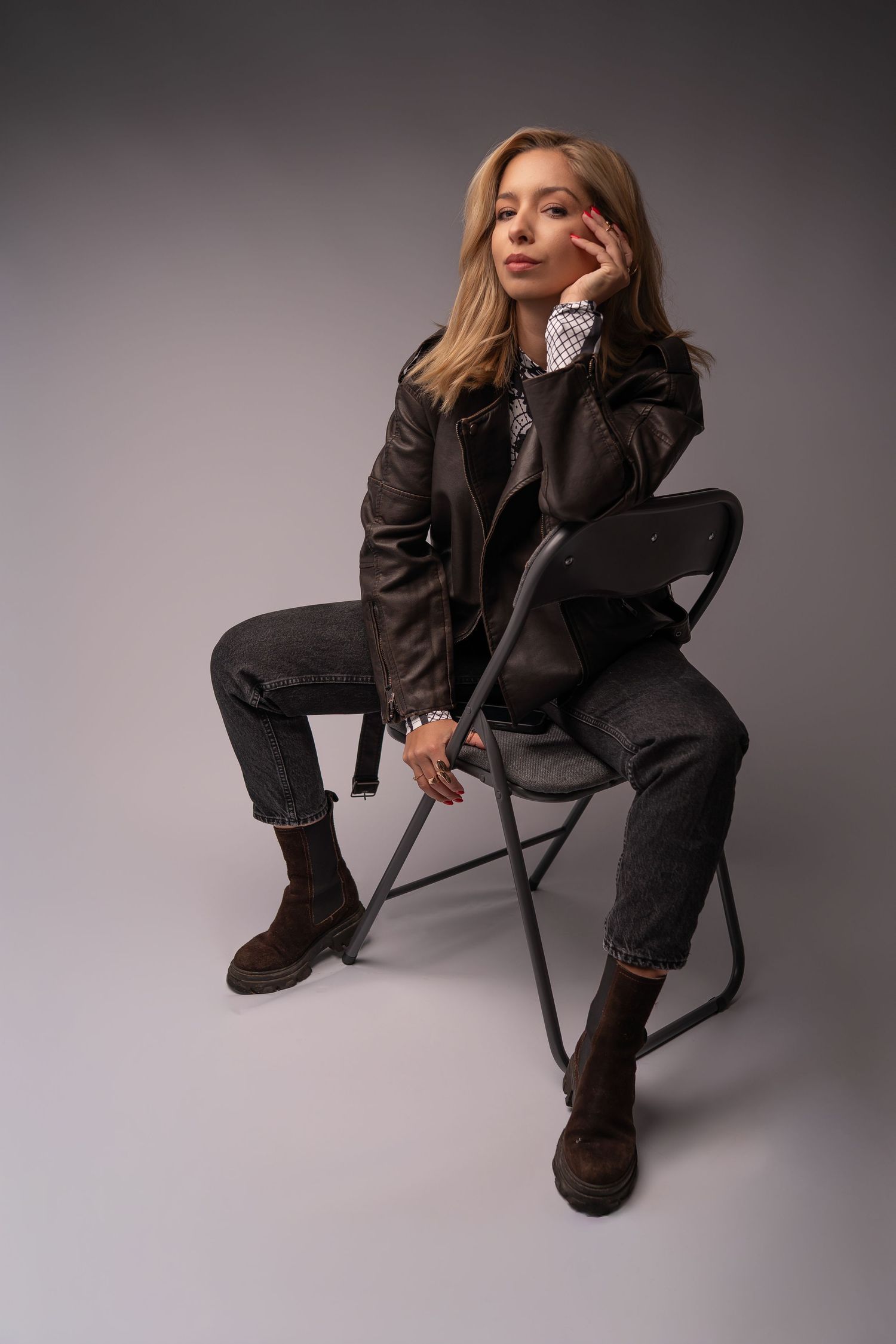 A person in a dark business suit and boots poses dramatically on a chair against a gray studio backdrop.