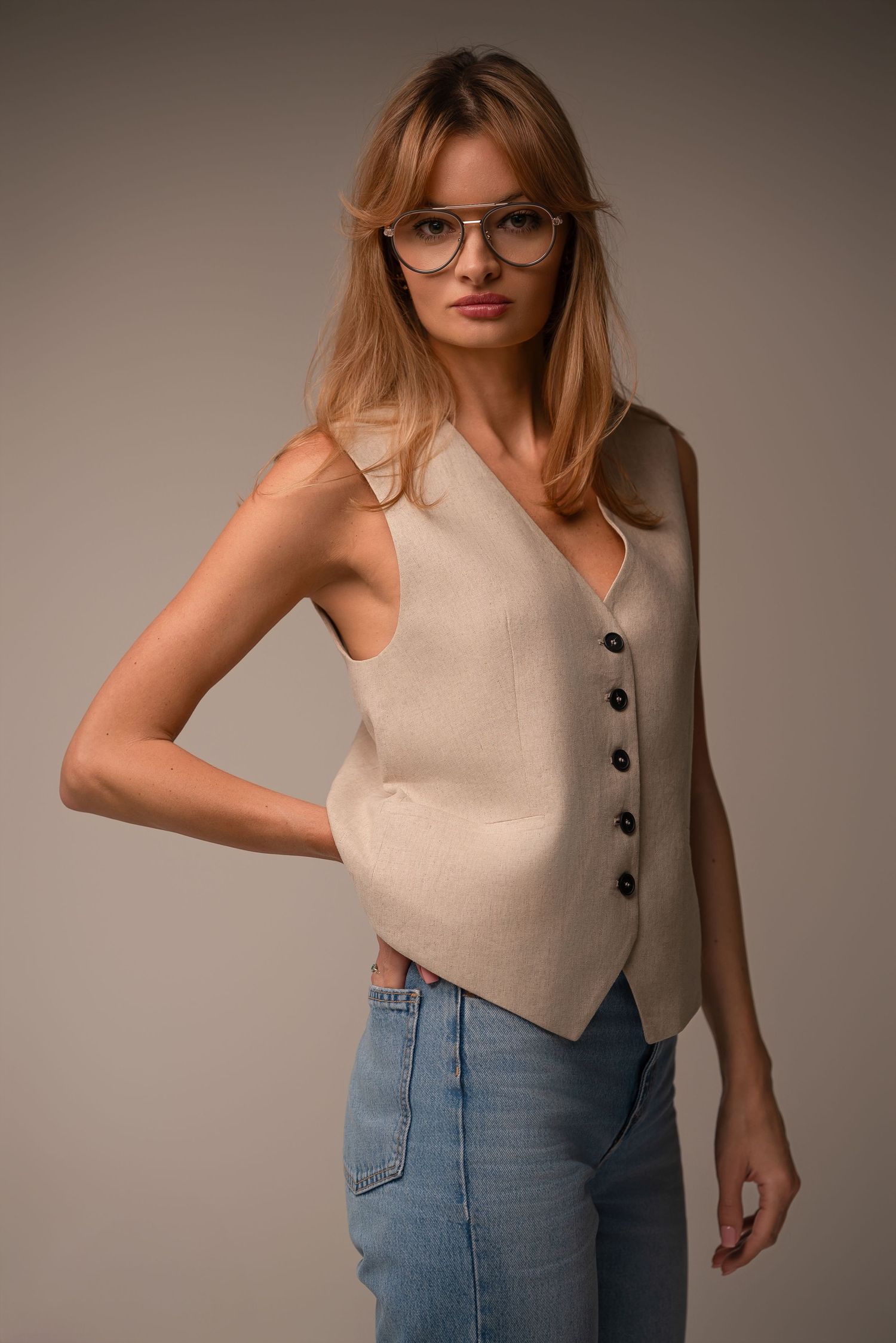 Model wearing beige button-up sleeveless vest with glasses and light blue jeans against neutral background.