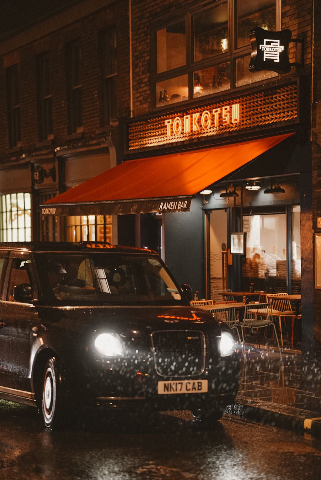 Black London taxi parked outside Tonkotsu restaurant with illuminated storefront on a rainy evening.