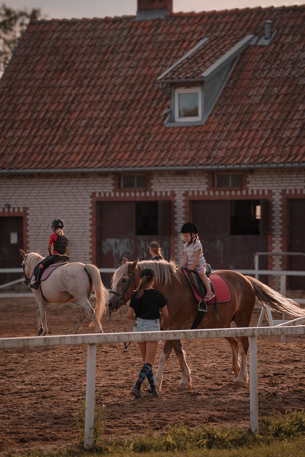 Equestrian scene at a stable with horses and riders in a sandy arena.