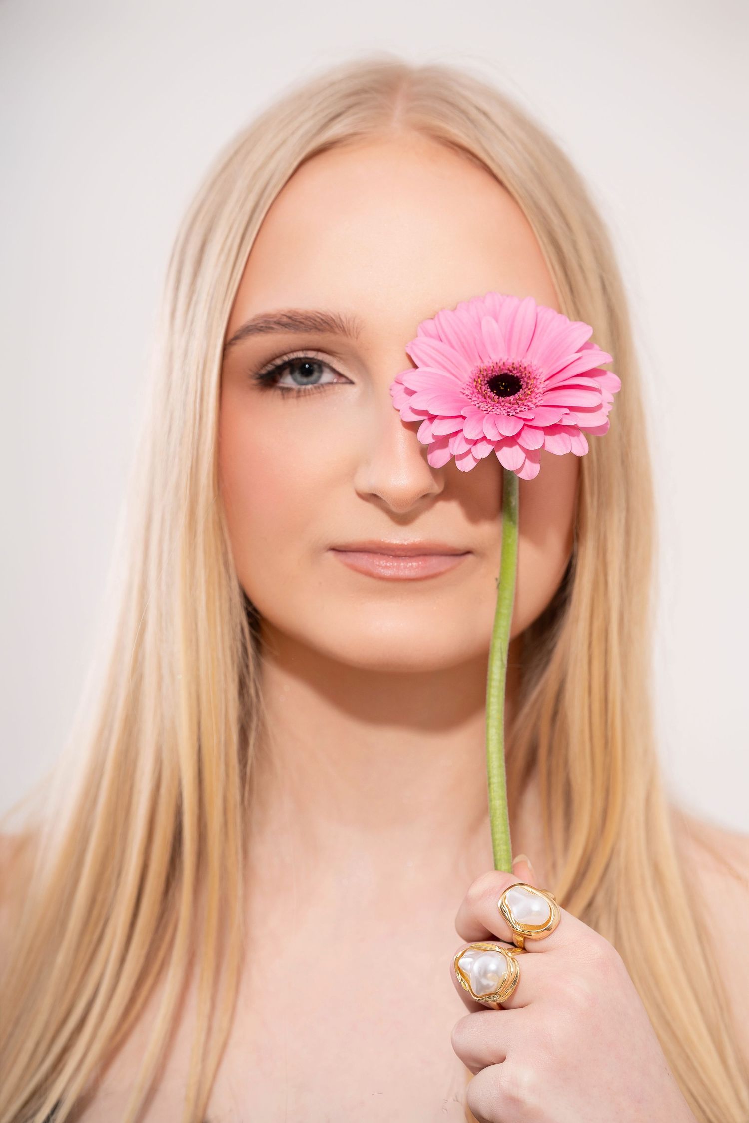 A pink gerbera daisy flower held in front of a face against a white background in a creative beauty portrait series.