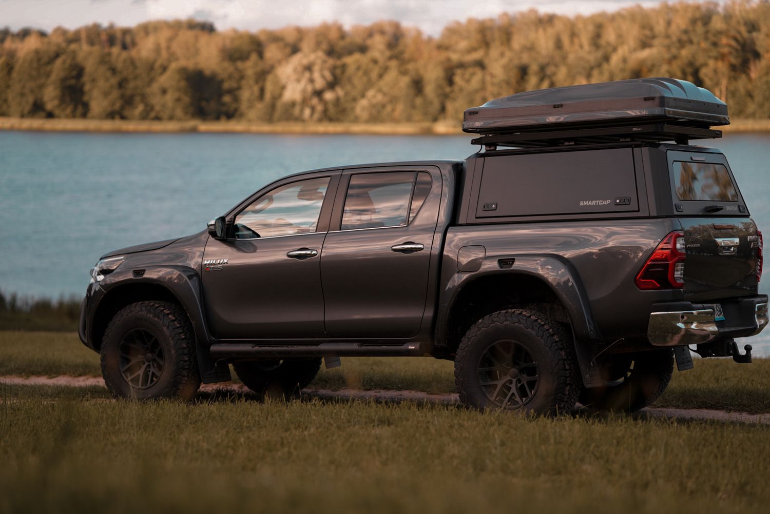 Black lifted pickup truck with roof cargo box parked beside a lake with forest in background.