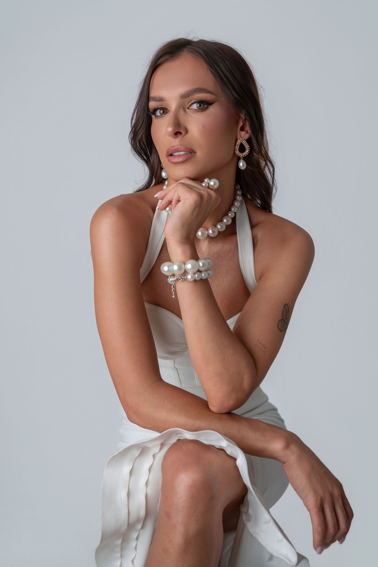Fashion model wearing white dress and pearl jewelry sits elegantly posed against light gray background.