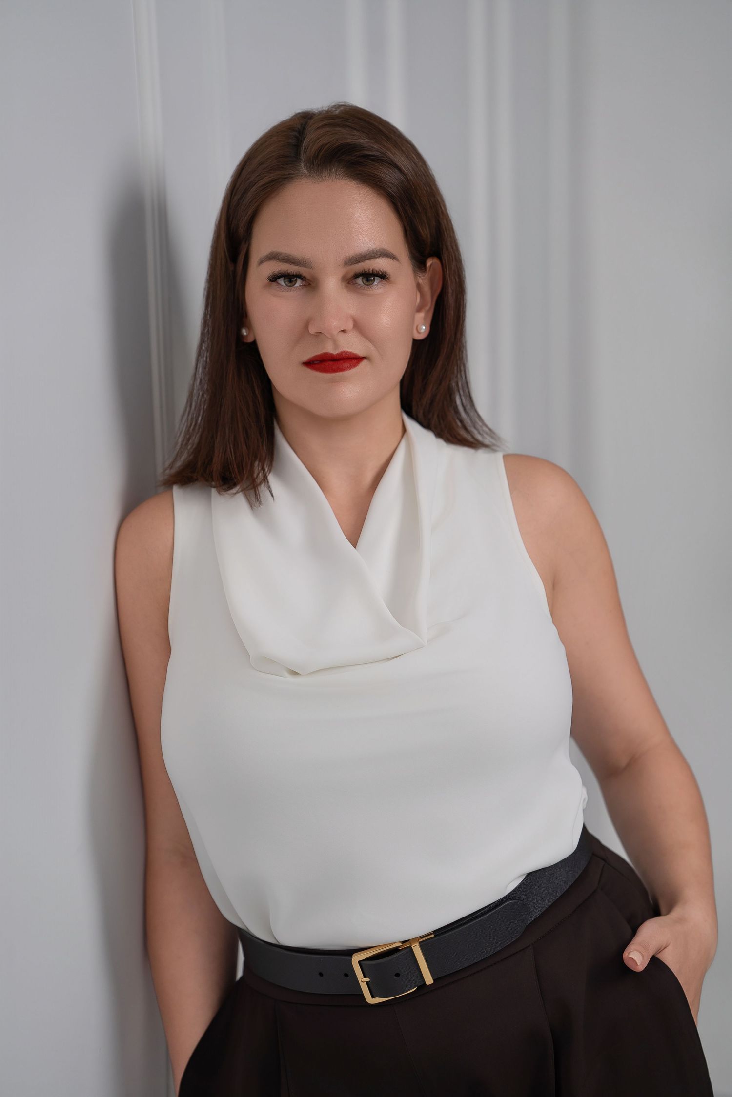 Professional studio portrait of a person wearing a sleeveless white cowl neck top with black skirt and belt against grey backdrop.