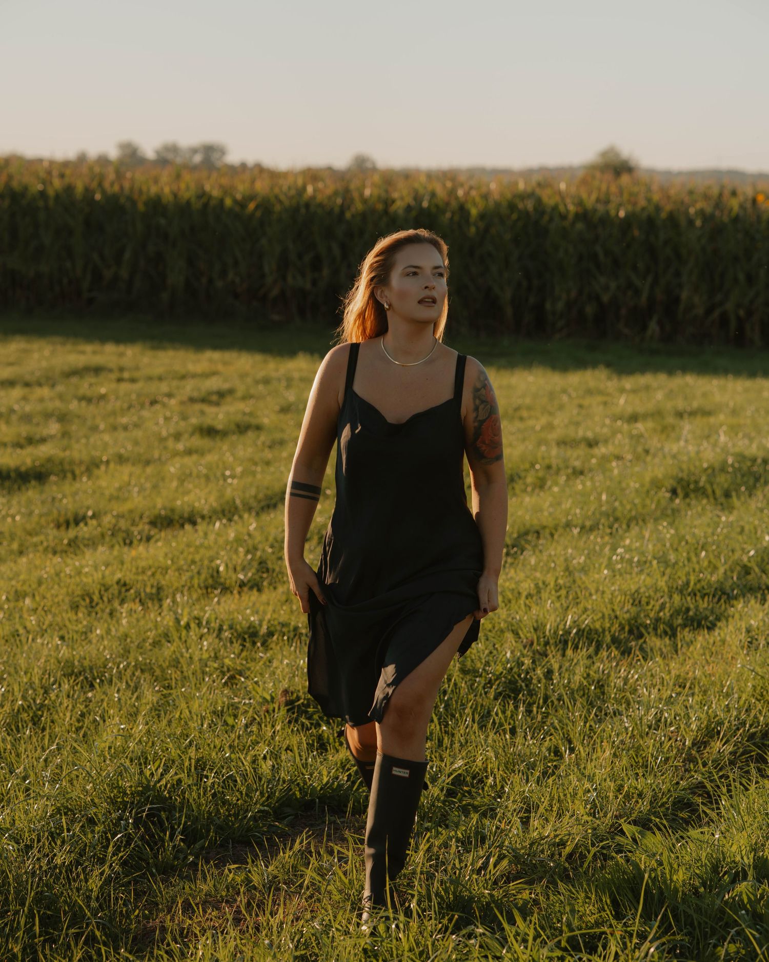 Figure in a black dress walks through a sunlit grassy meadow at golden hour with cornfields in the background.