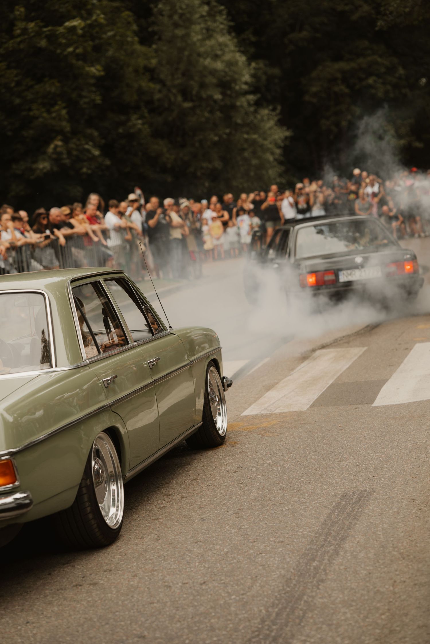 Vintage car performing burnout with white smoke as spectators watch from behind barriers at car show event.
