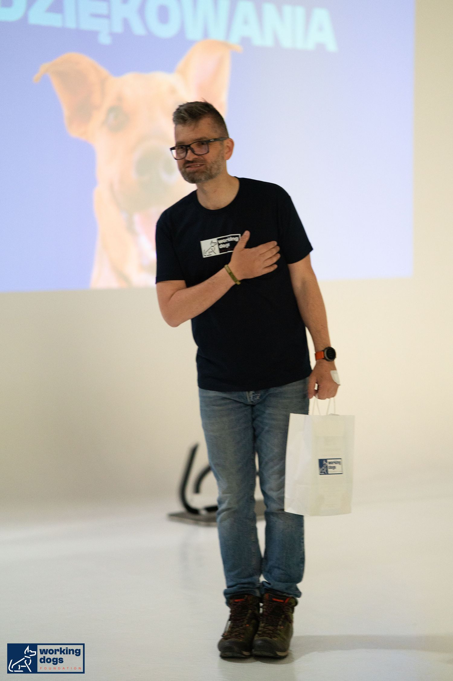 A presenter in a black t-shirt and jeans stands on stage with a projected pink animal silhouette in the background.