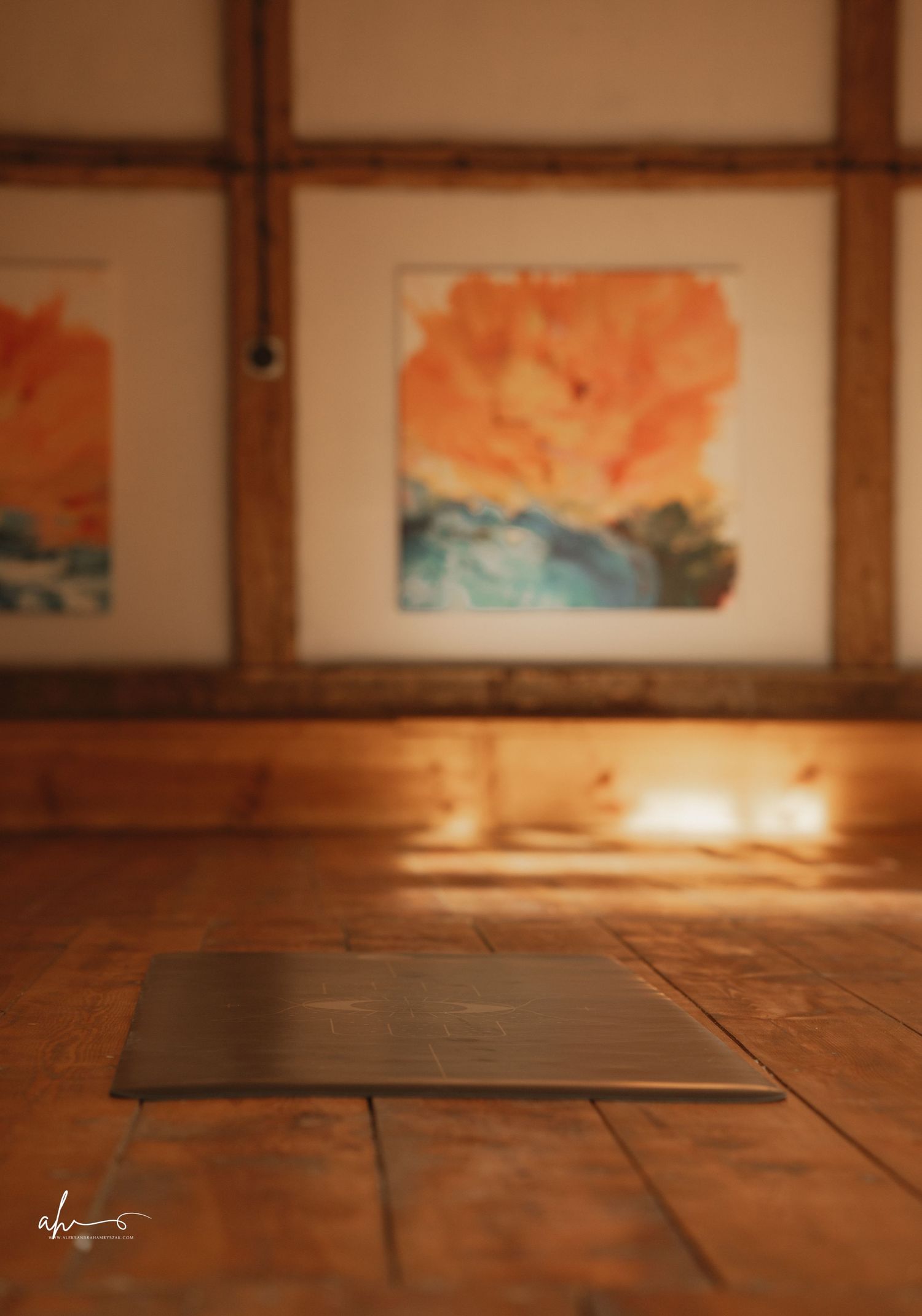 Warm candlelight illuminates an art gallery wall with orange watercolor paintings in wooden frames and a hardwood floor.