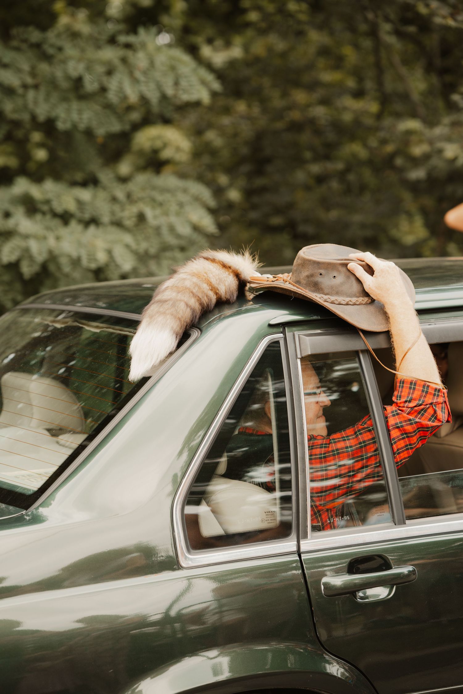 Someone wearing a red plaid shirt reaches out through a car window to grab a brown cowboy hat in a forest setting.