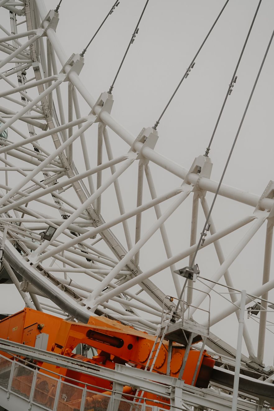 Close-up view of white structural support beams and cables on a Ferris wheel against an overcast sky.