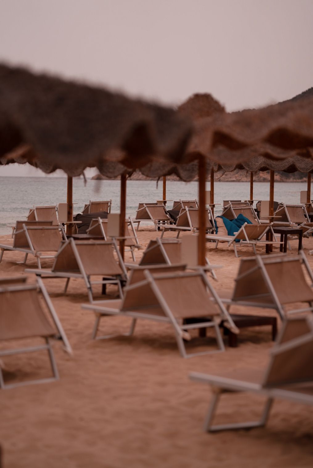 Empty beach lounge chairs with thatched umbrellas arranged on sandy shore at sunset.