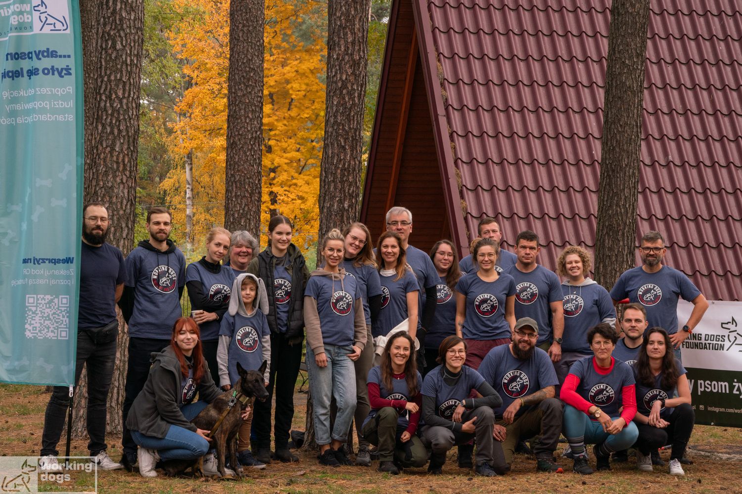 Large group wearing matching navy shirts poses for outdoor photo in front of autumn trees.