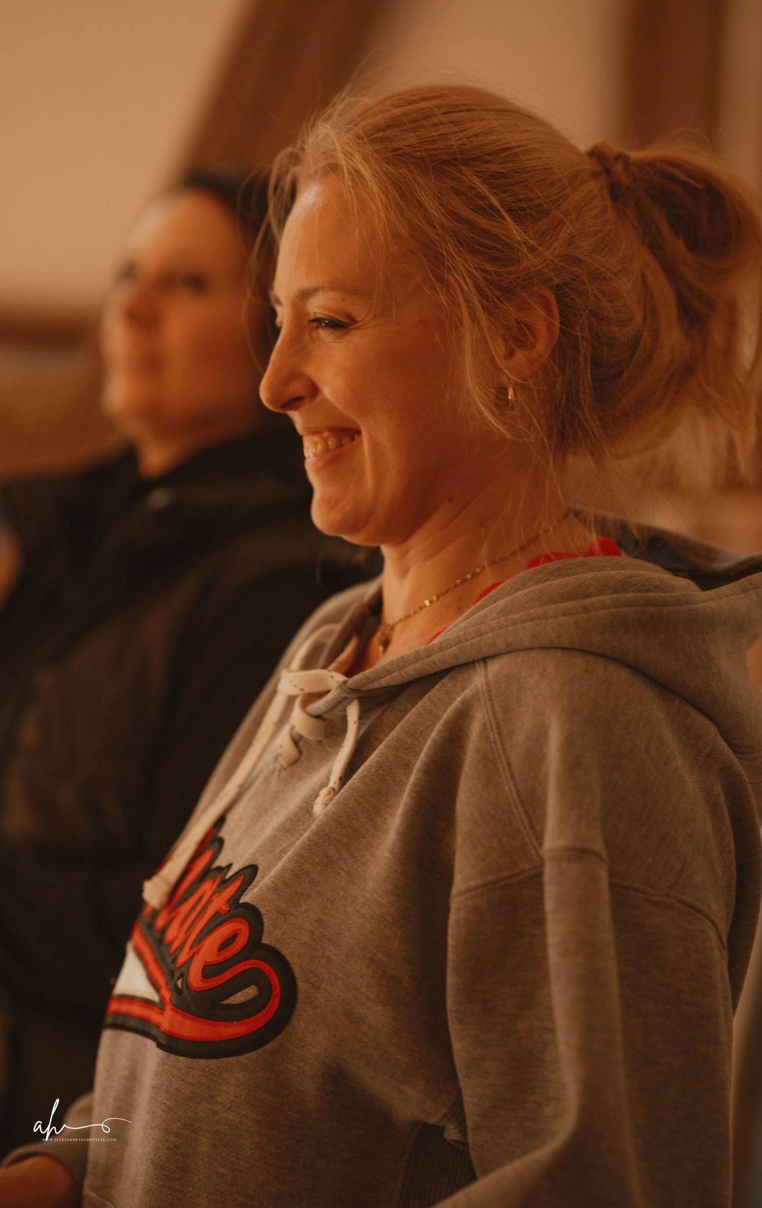 Someone smiling warmly while wearing a gray Nike hoodie in warm lighting with a blurred background.