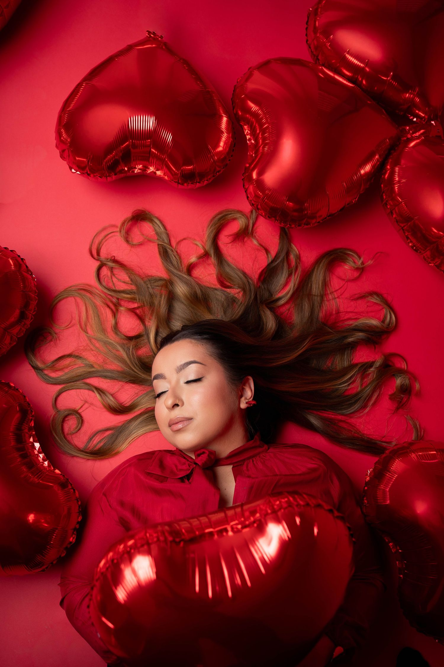 A dreamy Valentine's themed photoshoot with red heart balloons and flowing hair against a crimson background.