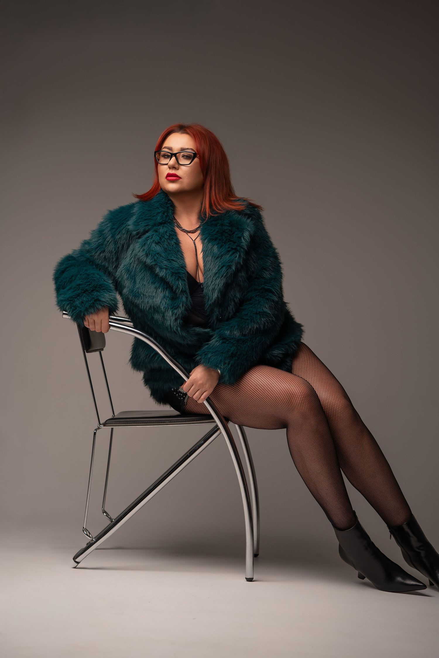 A model in a teal fur coat and black stockings poses dramatically on a modern chrome chair against a gray backdrop.
