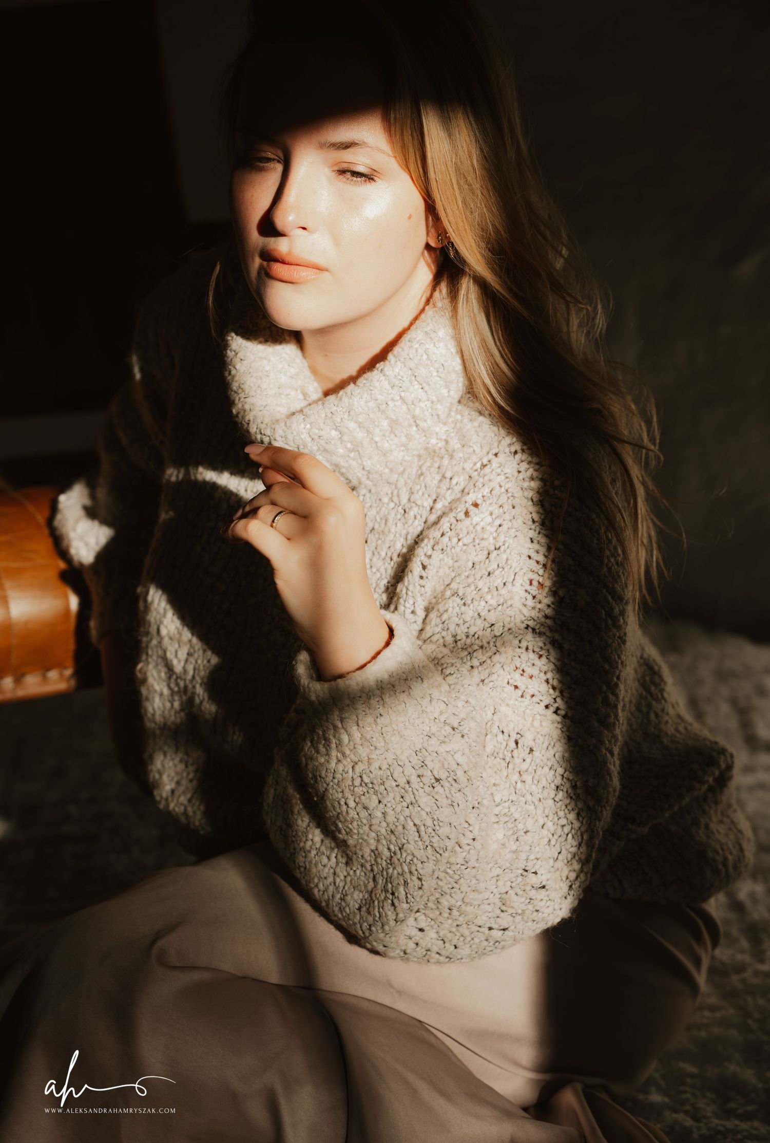 Artistic portrait in moody lighting showing someone in cozy white sweater sitting in relaxed pose with soft lighting.