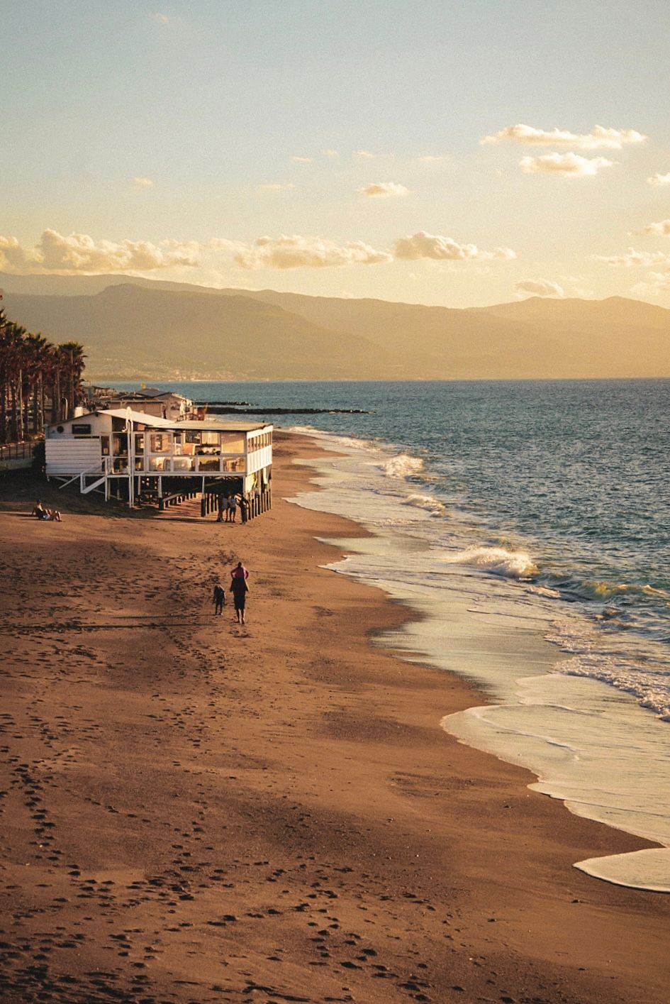 A beachfront establishment sits on stilts along a sandy shoreline at golden hour with mountains in the distance.