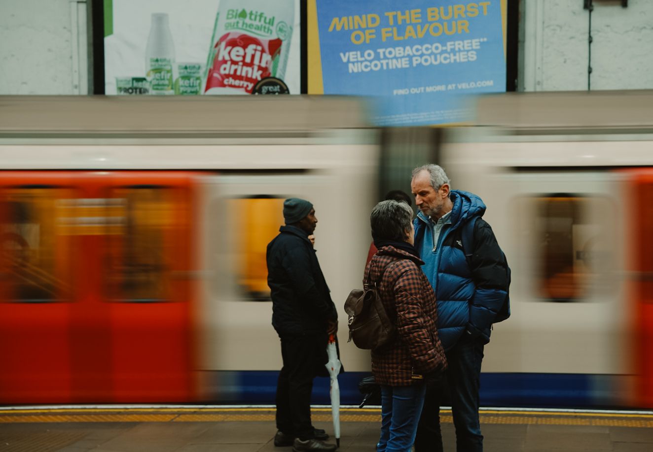 People wait on a subway platform as a red train blurs past in motion at an underground London tube station.