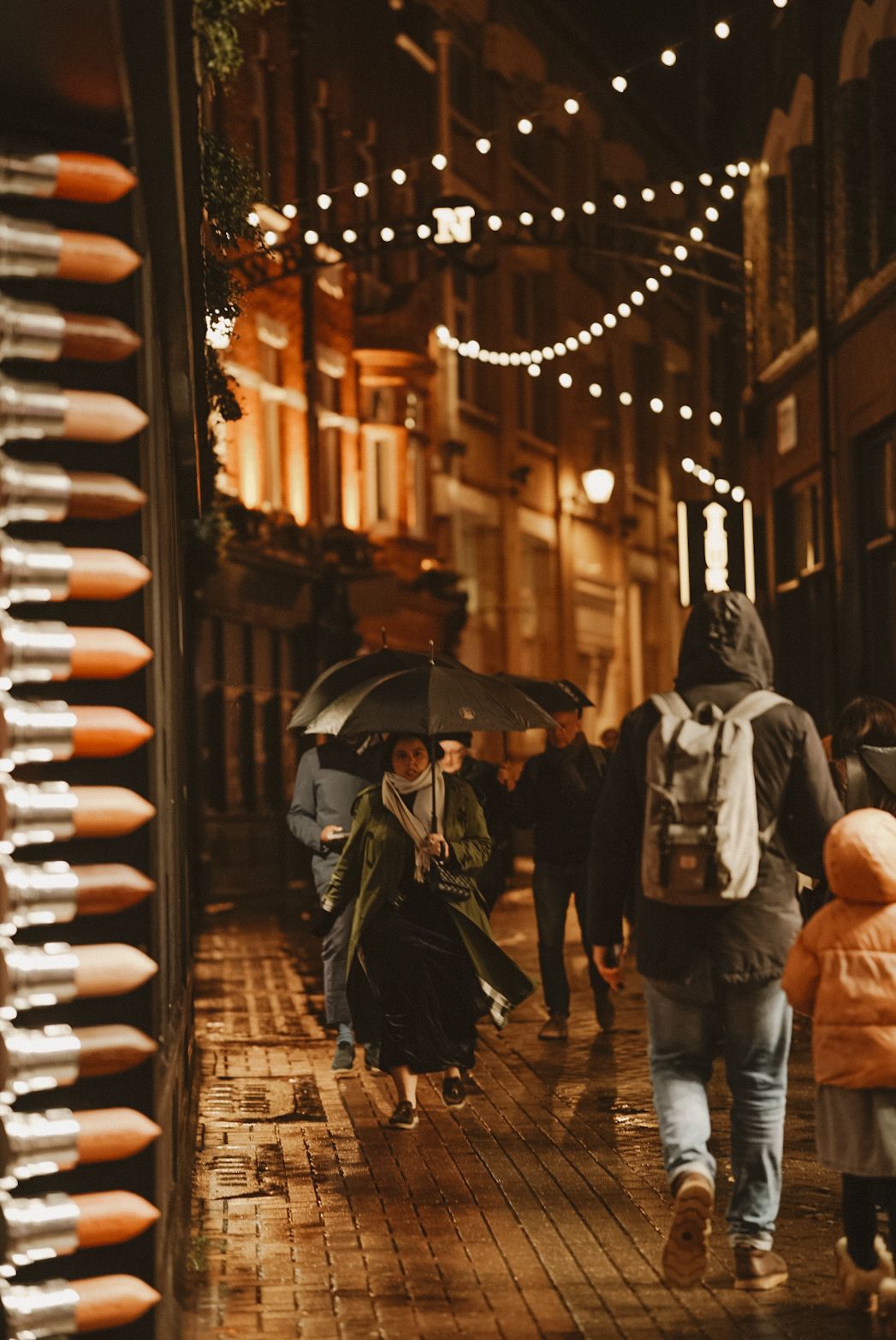 A rainy evening city street with string lights overhead and pedestrians walking with umbrellas on wet cobblestones.