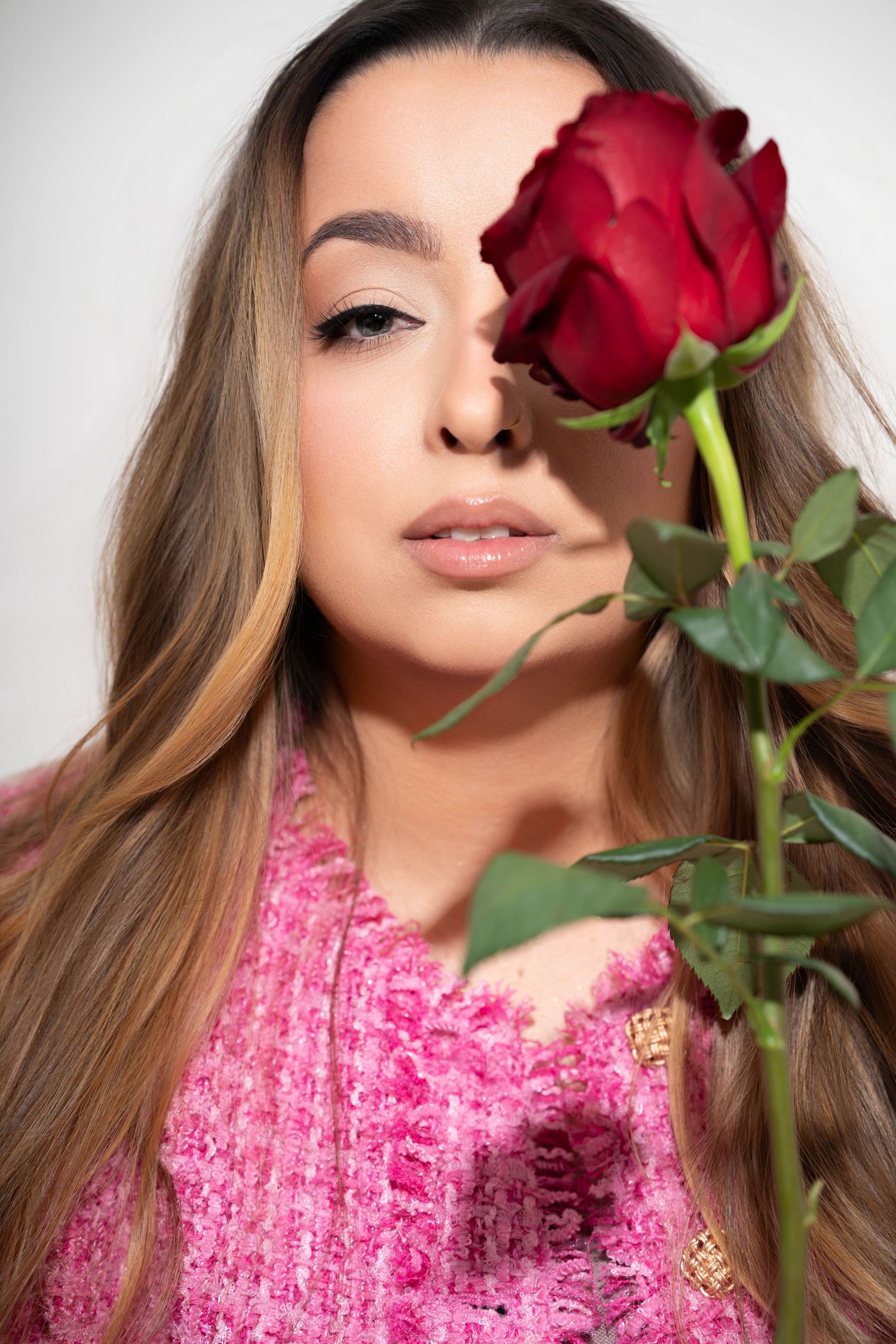 A red rose stem partially covers a portrait of someone wearing a pink textured top with long wavy brown hair.