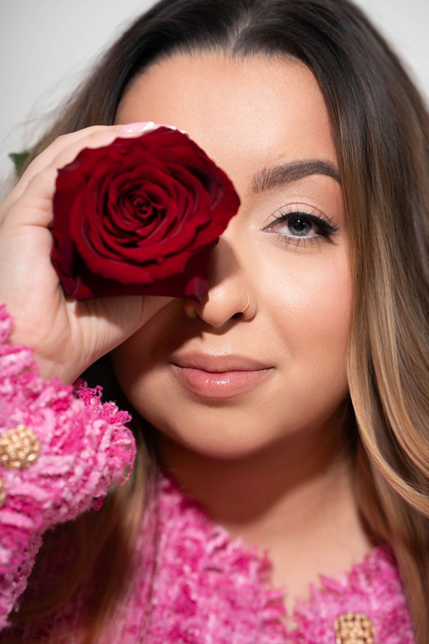 A deep red rose held up to cover one eye, with pink floral attire and soft makeup creating a romantic beauty portrait.