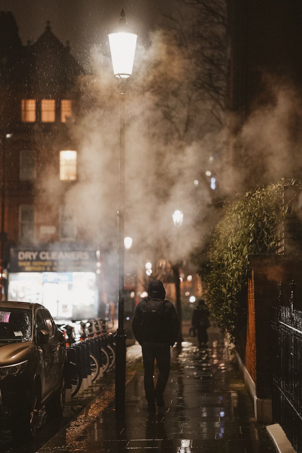 Foggy night street scene with glowing lampposts and a dry cleaners shop sign visible in the background.