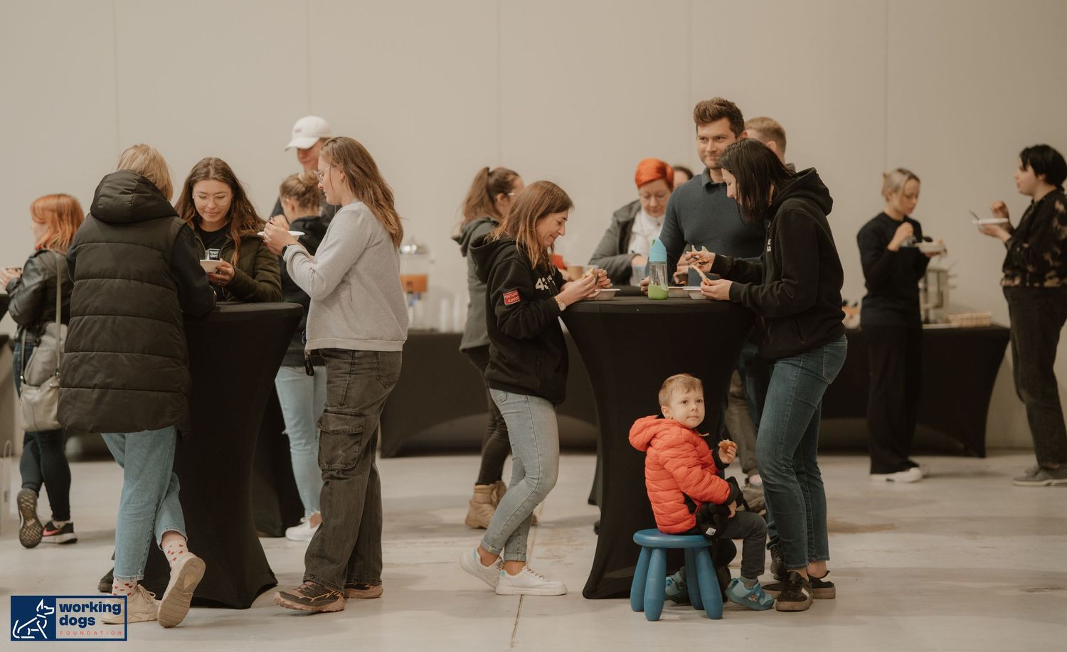 People gathered at an indoor event space with a small child on a blue chair in the foreground.