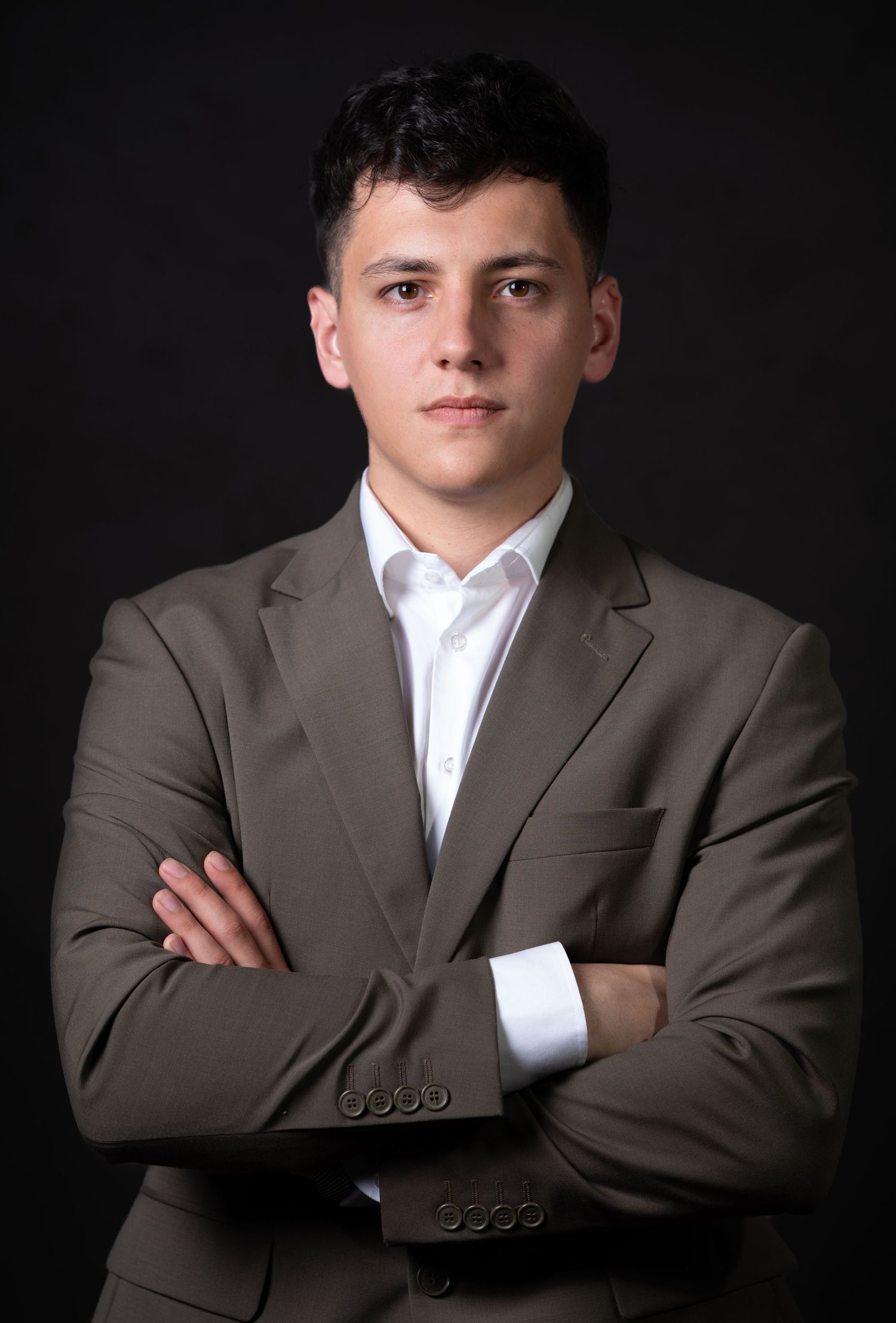 Professional studio portrait of a young professional in a brown suit and white shirt with arms crossed against dark background.