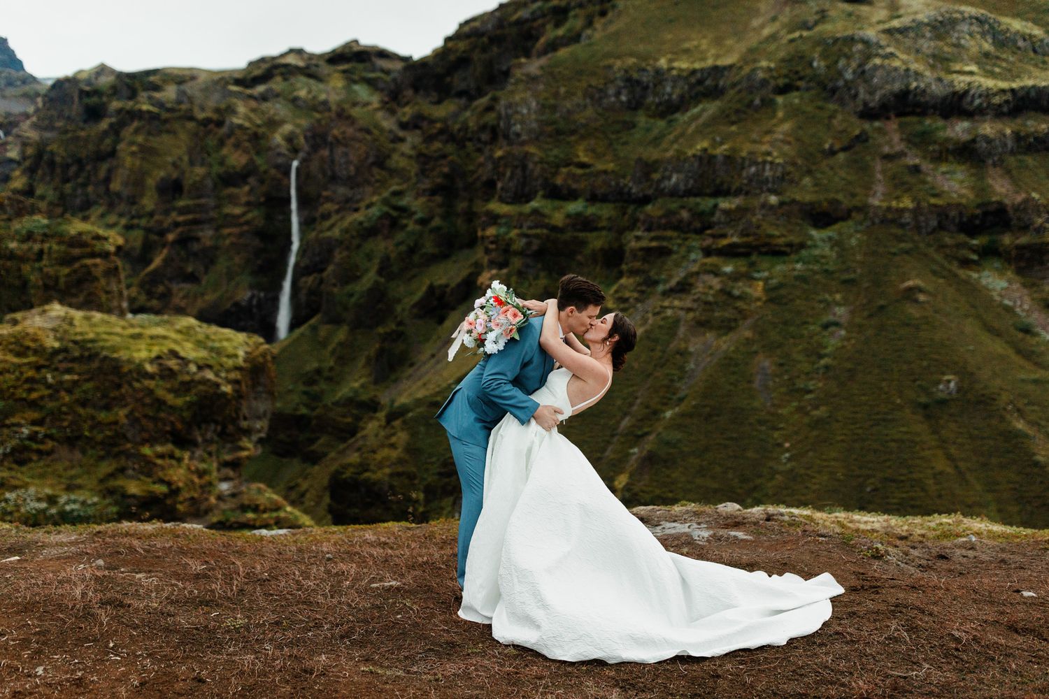 A couple in wedding attire poses on a dramatic mountain cliff with a cascading waterfall in the background.