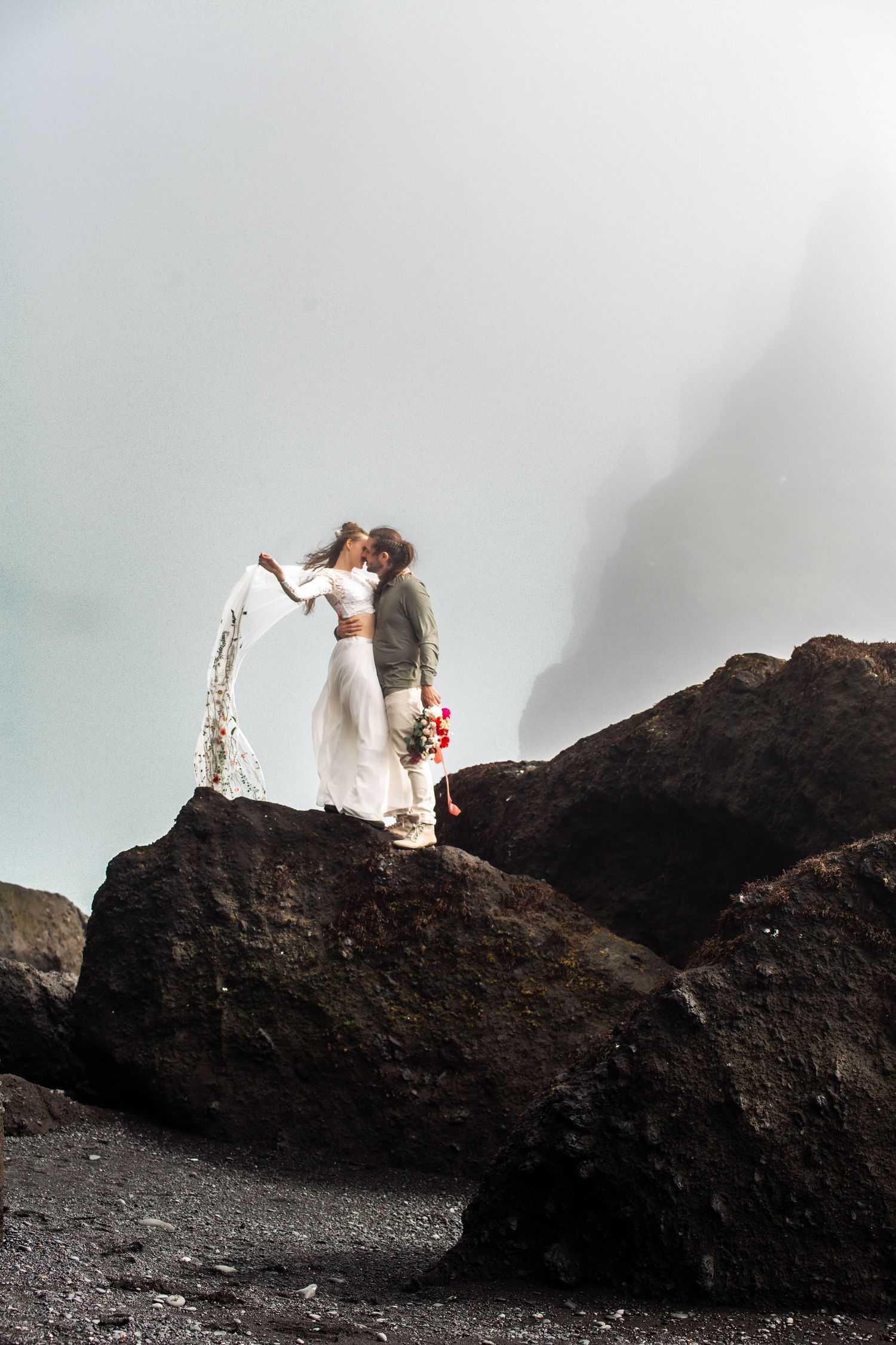 A romantic wedding couple stands on dramatic black volcanic rocks with flowing white dress and veil against misty sky.