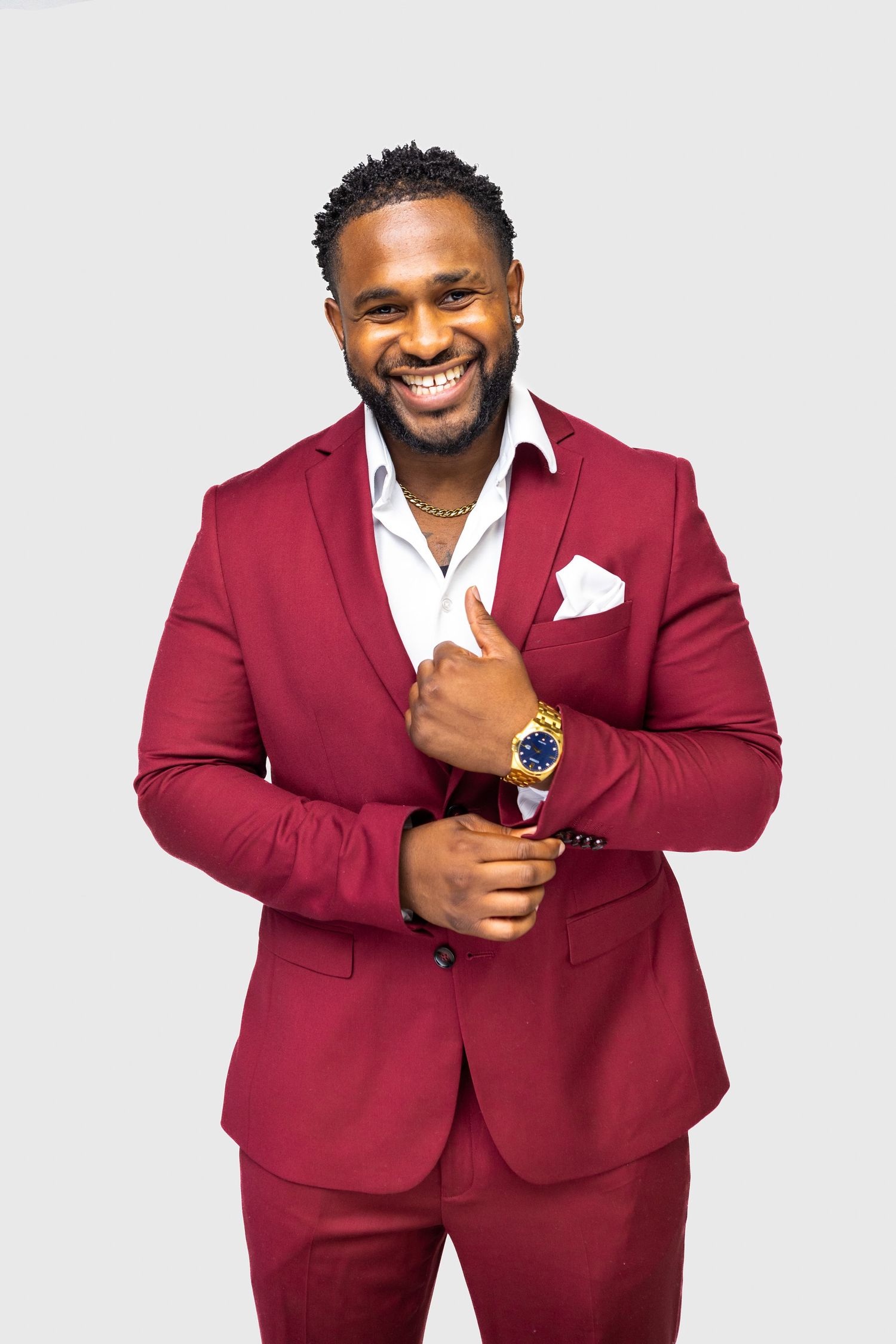 A person wearing a burgundy suit and white dress shirt smiles while adjusting their watch against a light gray background.