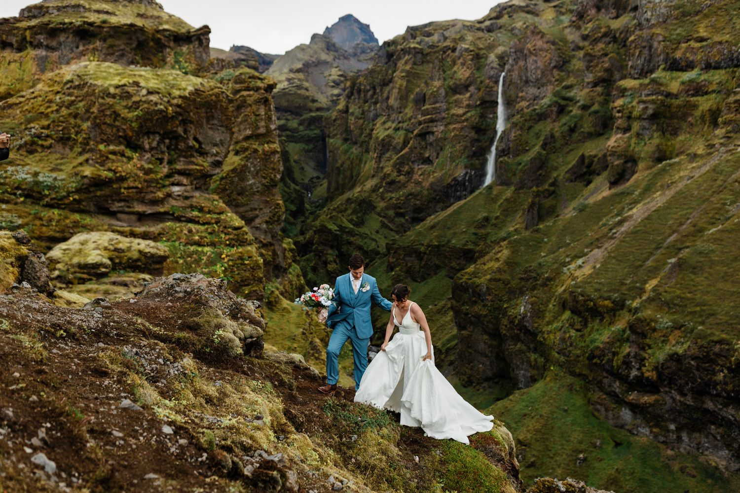 A couple in wedding attire stands near a dramatic waterfall in Iceland's mountainous landscape.