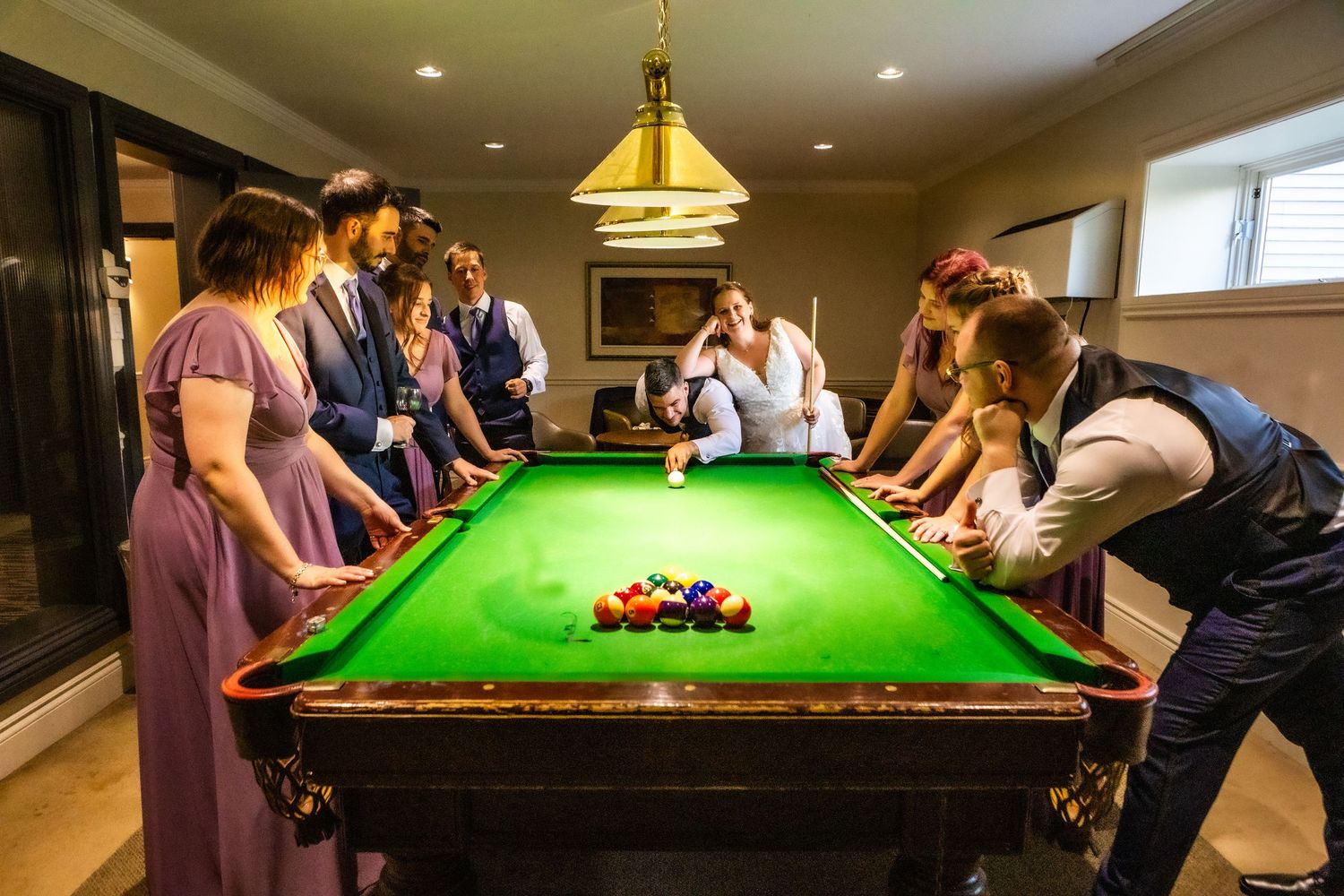People gather around a green snooker table during a social game in a warmly lit room.