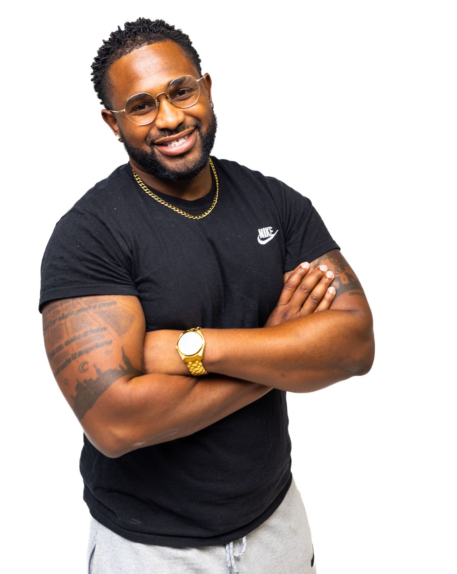 Athletic man in black t-shirt and grey shorts poses with crossed arms and bright smile against white background.