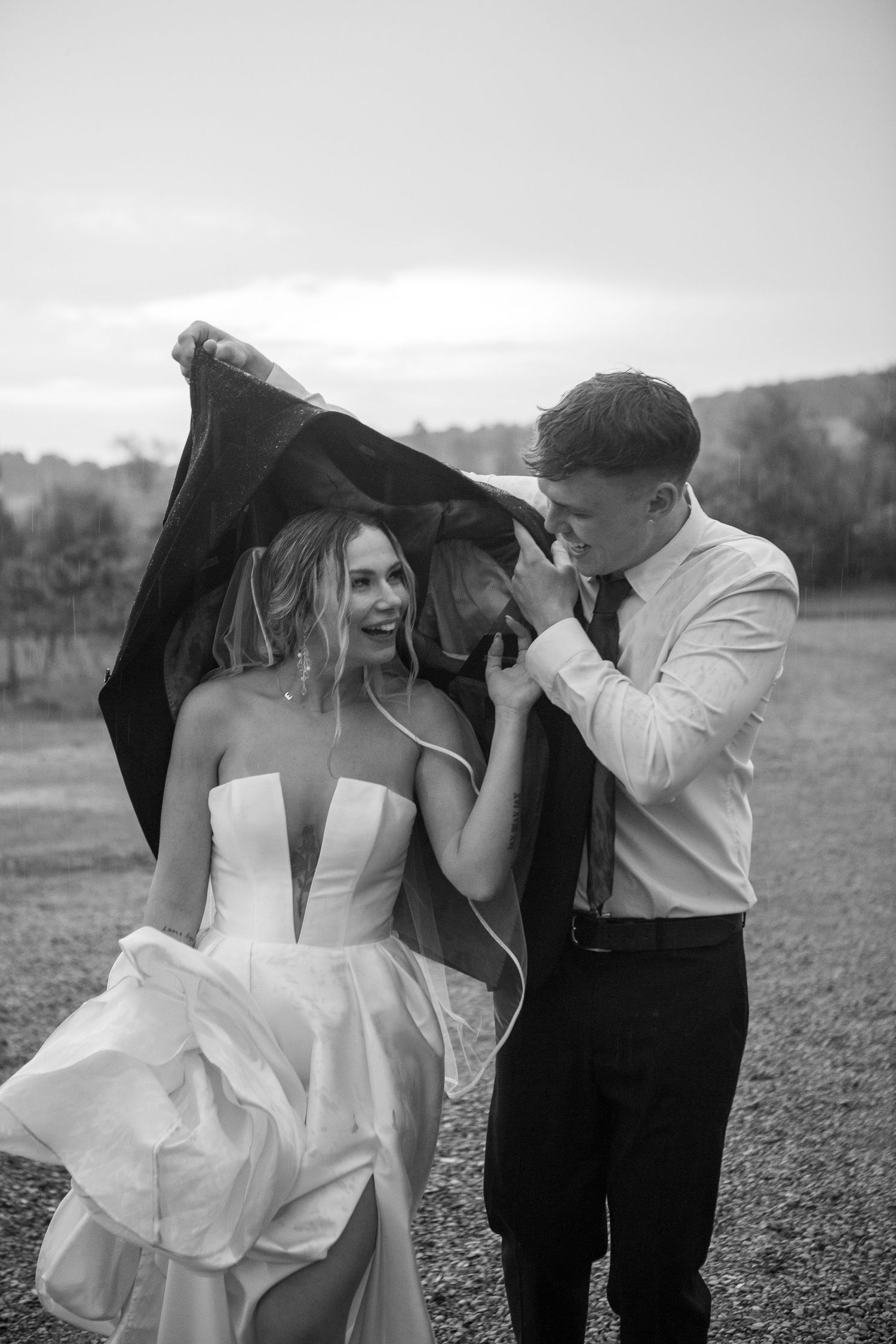 Black and white photo of couple sharing intimate moment under jacket in outdoor setting.