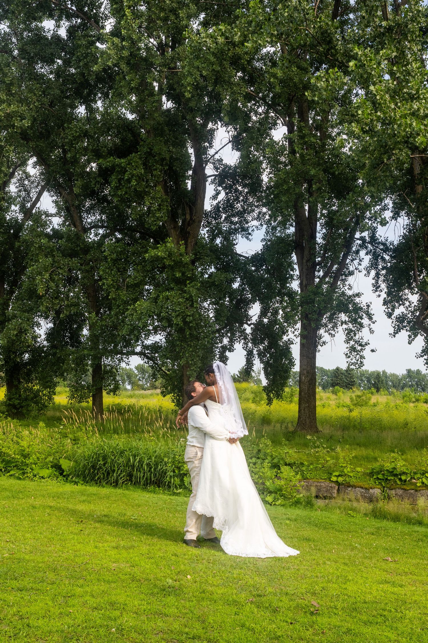 A couple in wedding attire walks together across a green hillside with tall trees in the background.