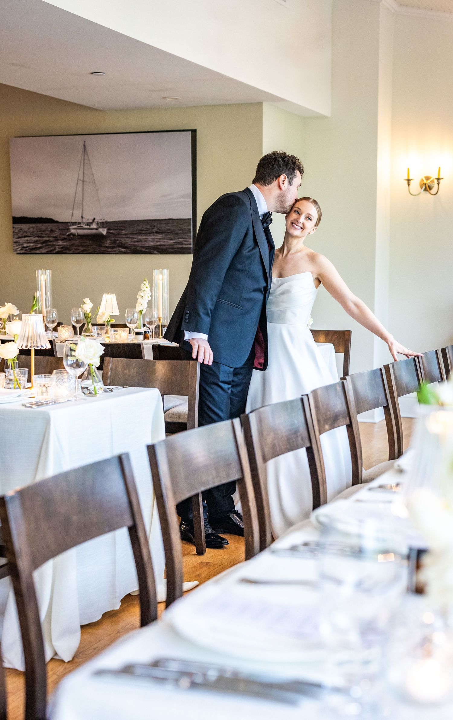 A wedding reception setup with elegant long tables, white linens, and dark wooden chairs in a bright indoor venue.