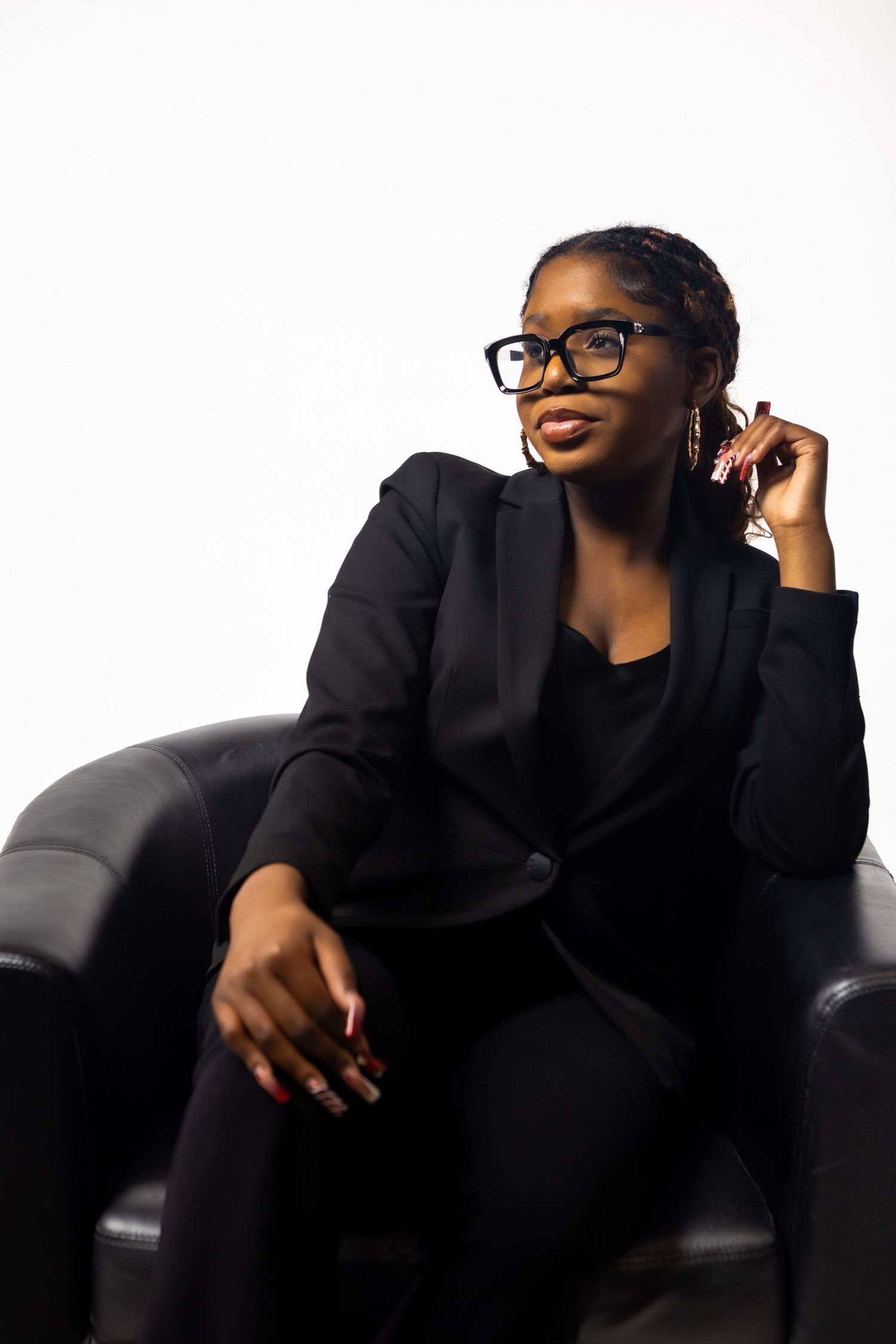 Professional wearing black attire and glasses poses on leather couch in artistic portrait series.