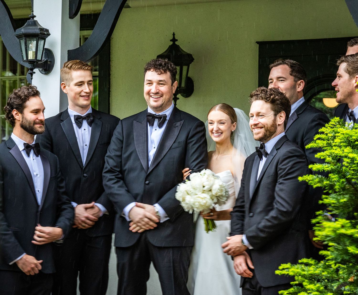 Wedding party stands in formal attire on steps with lush green foliage in the background.