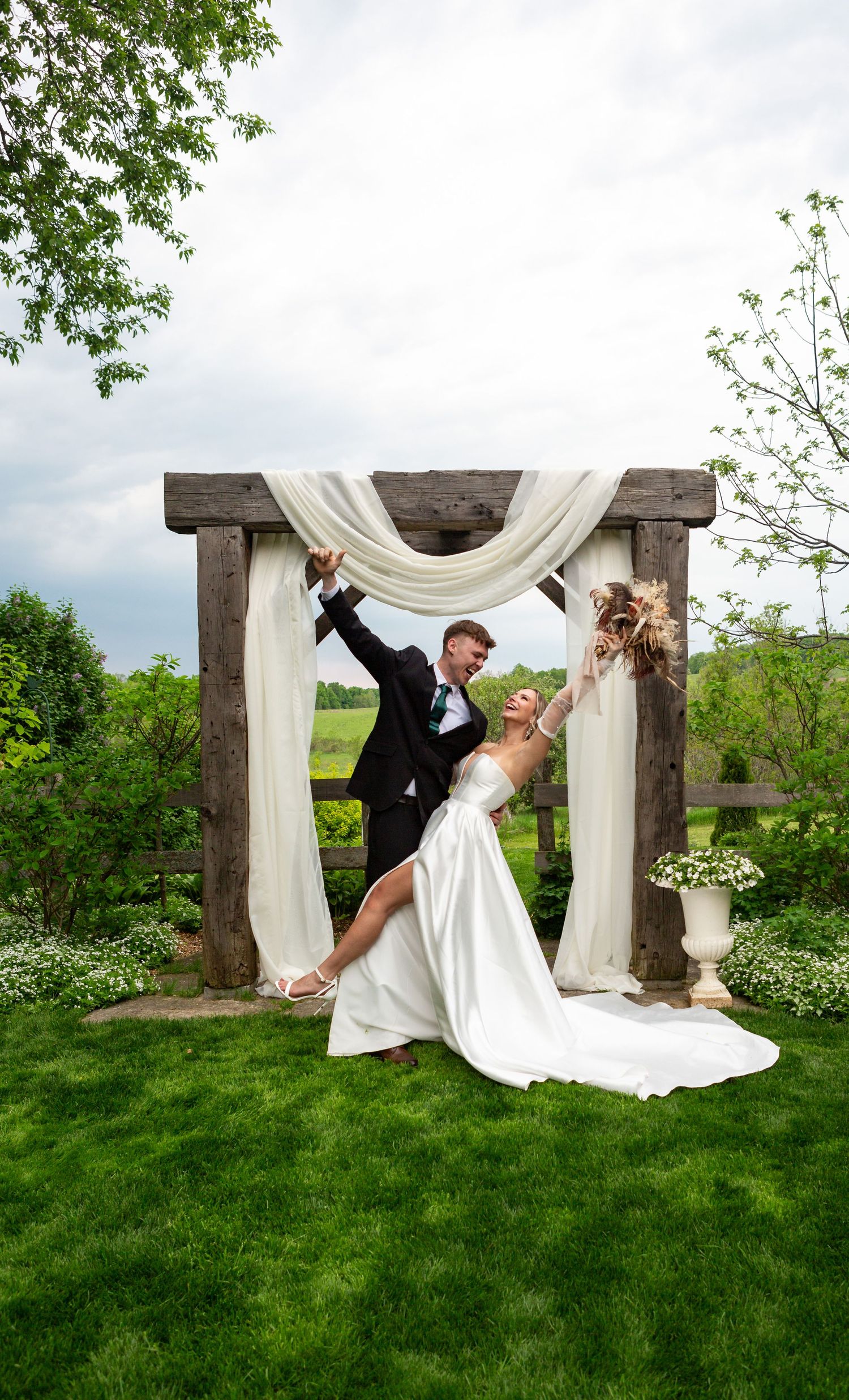 Newlyweds strike playful pose under wooden arch decorated with white fabric.