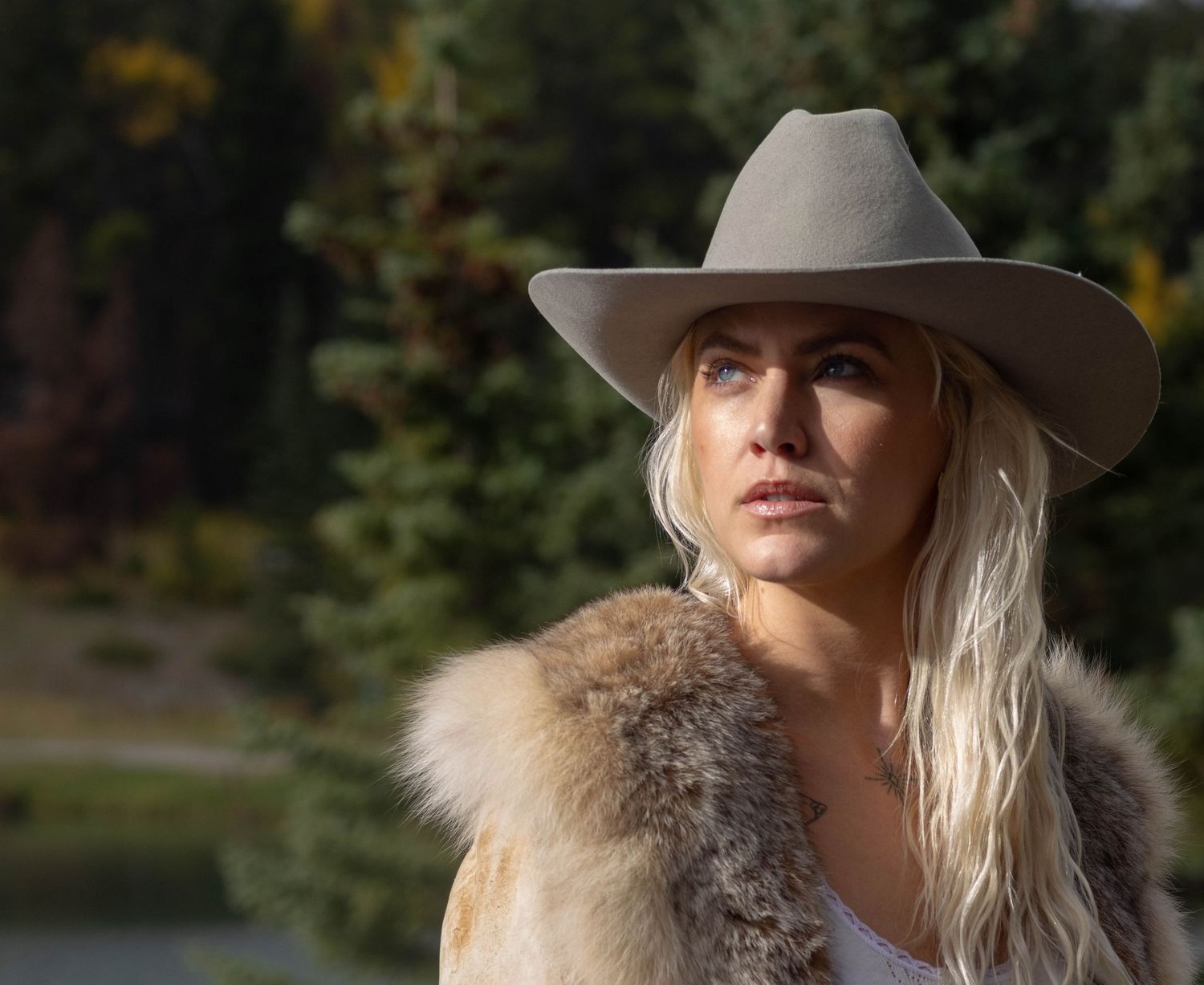 A person wearing a cowboy hat and fur coat poses outdoors against a natural background in autumn.