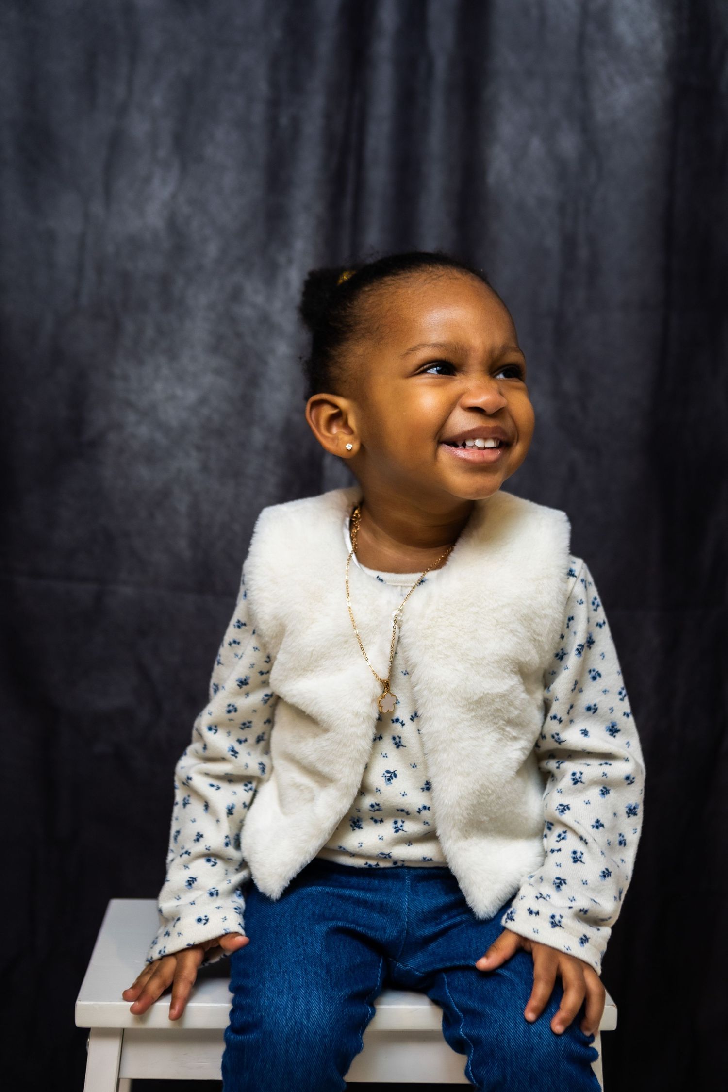 A young child wearing a white furry vest and floral shirt sits on a white stool against a dark background, smiling brightly.
