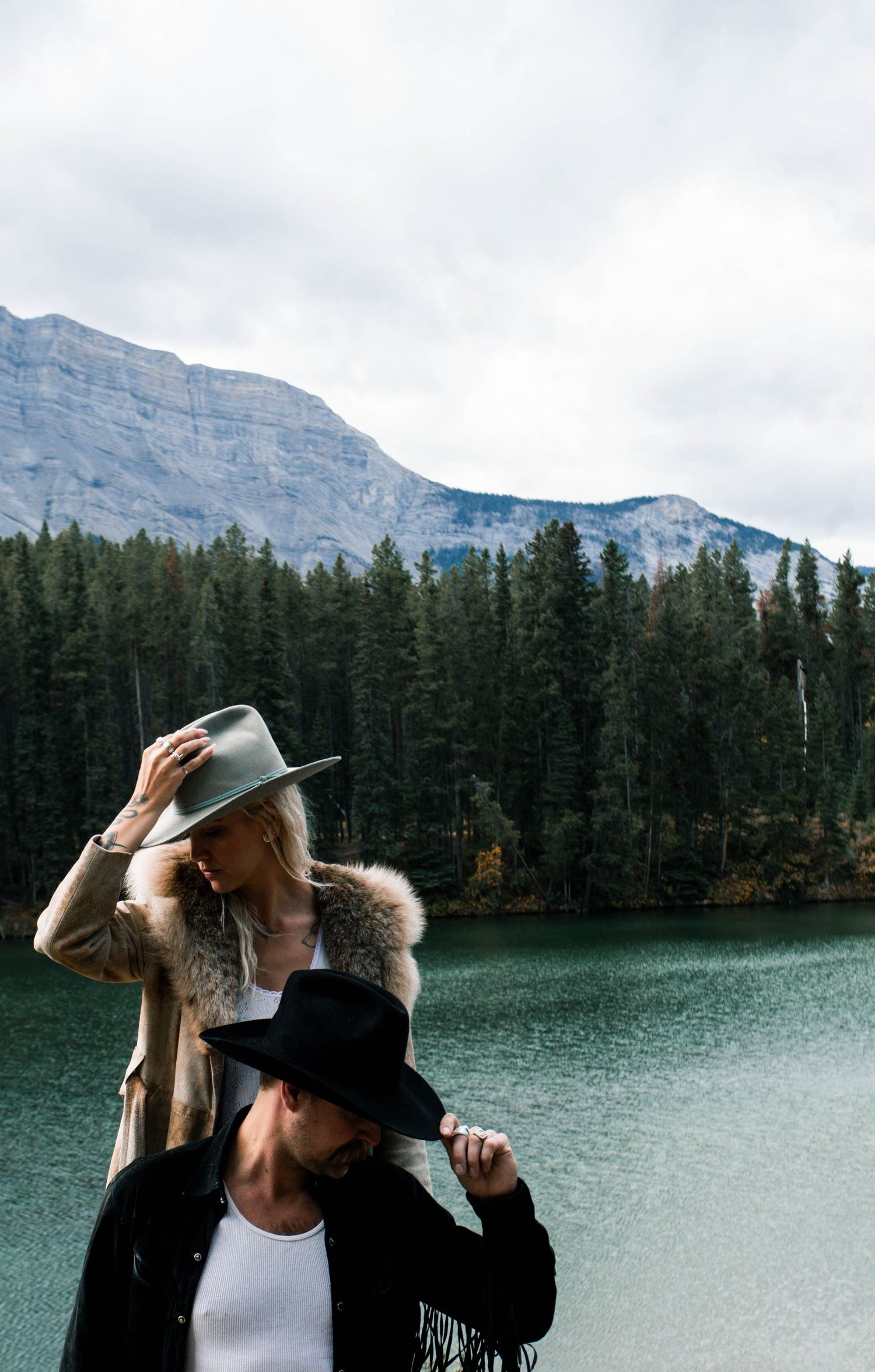 Two people in hats pose by a turquoise mountain lake surrounded by evergreen forest and rocky peaks.