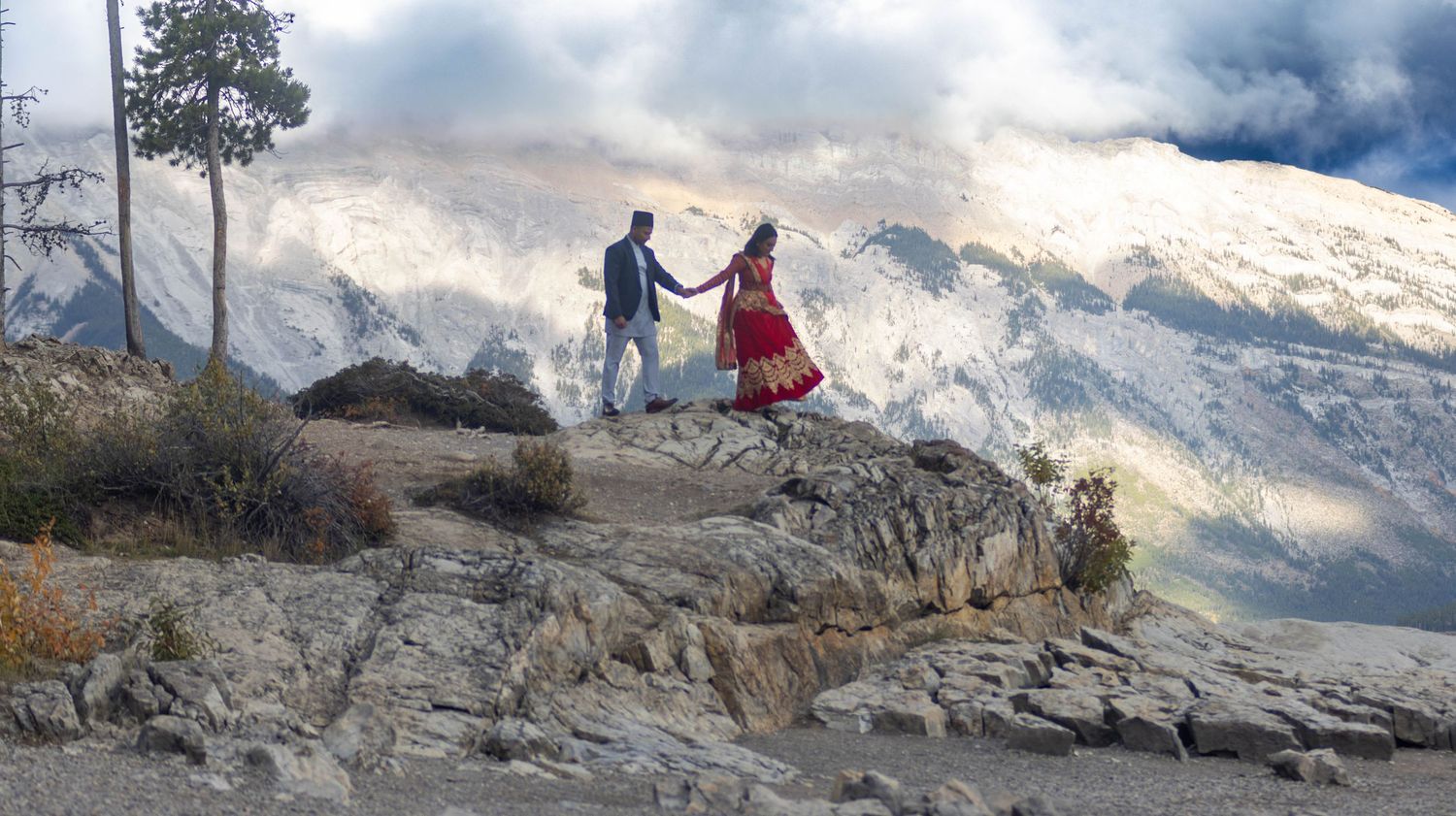 Panoramic mountain landscape with rocky terrain and snow-capped peaks against a dramatic cloudy sky.
