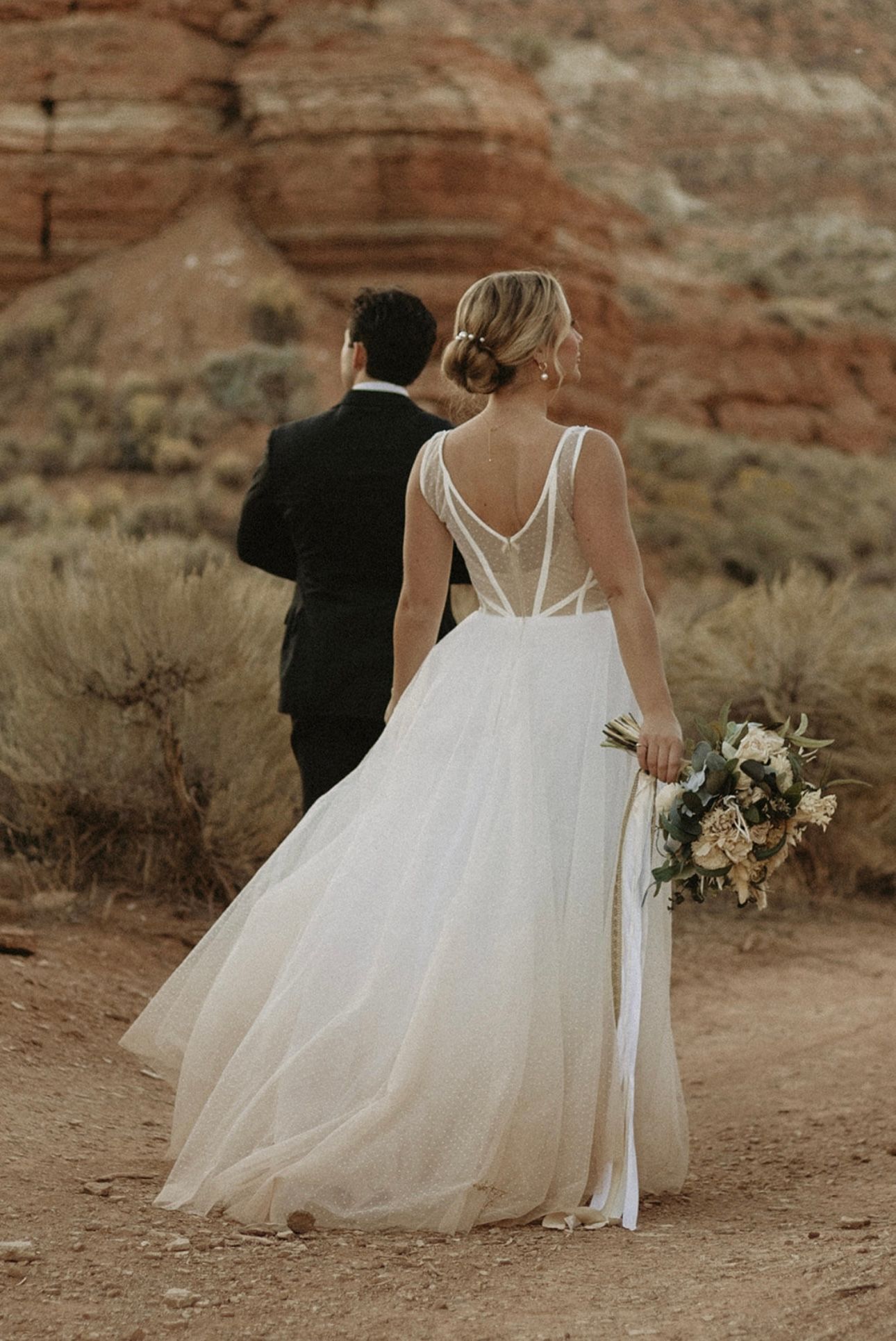 Bride in white flowing dress with groom walking along desert red rock landscape.