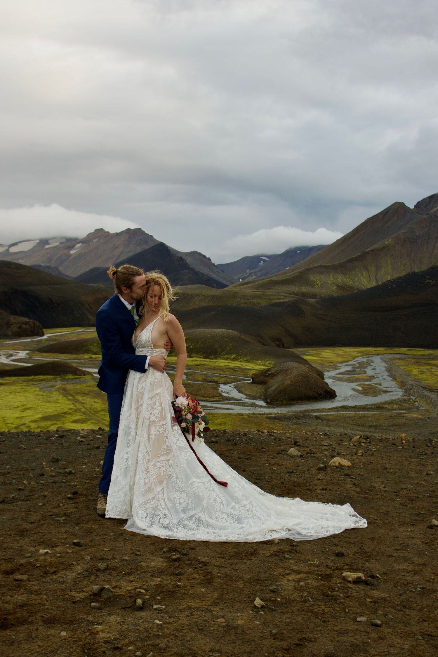 A couple in wedding attire embraces on a dramatic Icelandic landscape with winding rivers and mountains in the background.