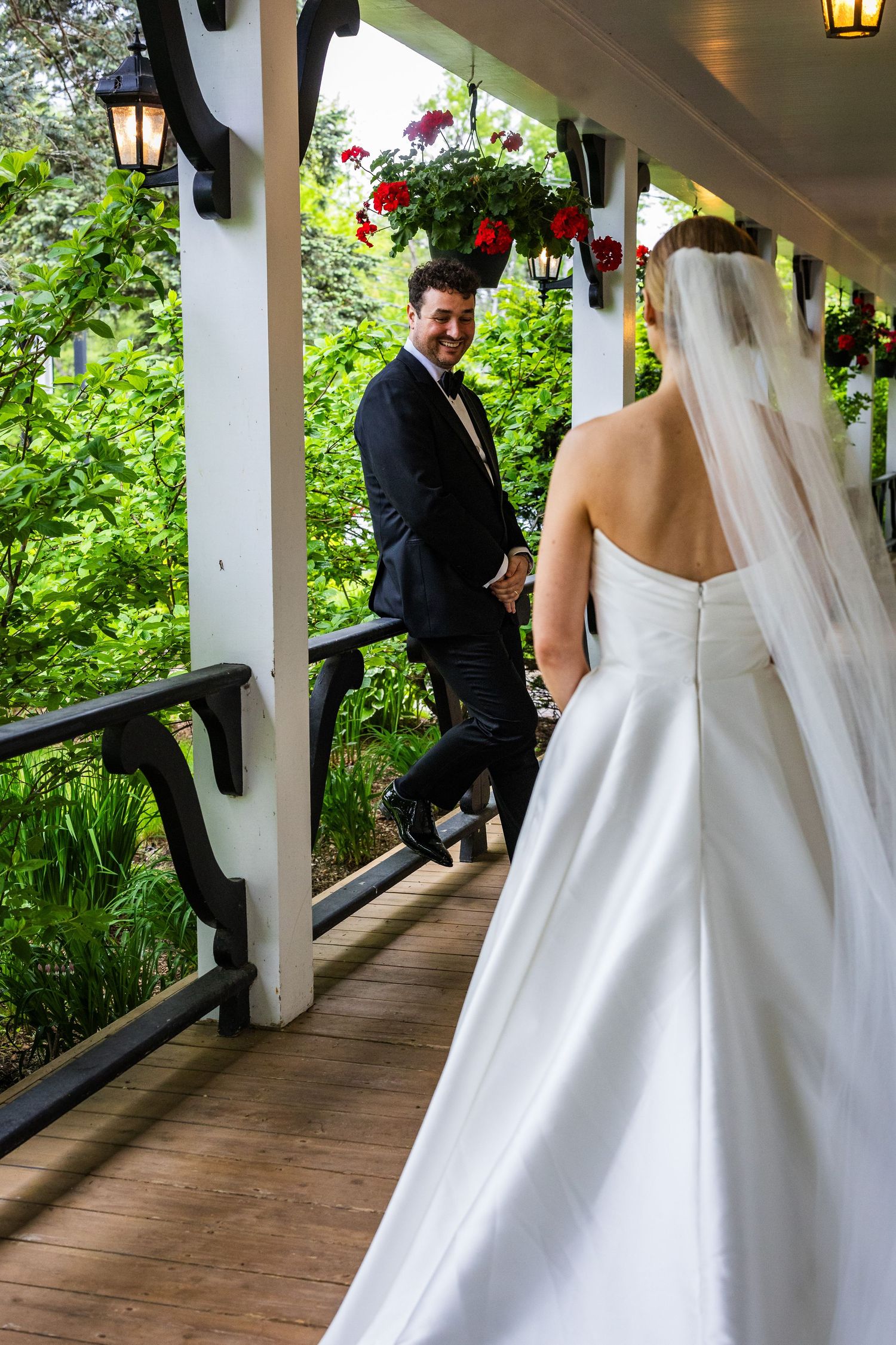 A bride and groom walk along a covered wooden porch with lanterns and greenery creating a romantic atmosphere.