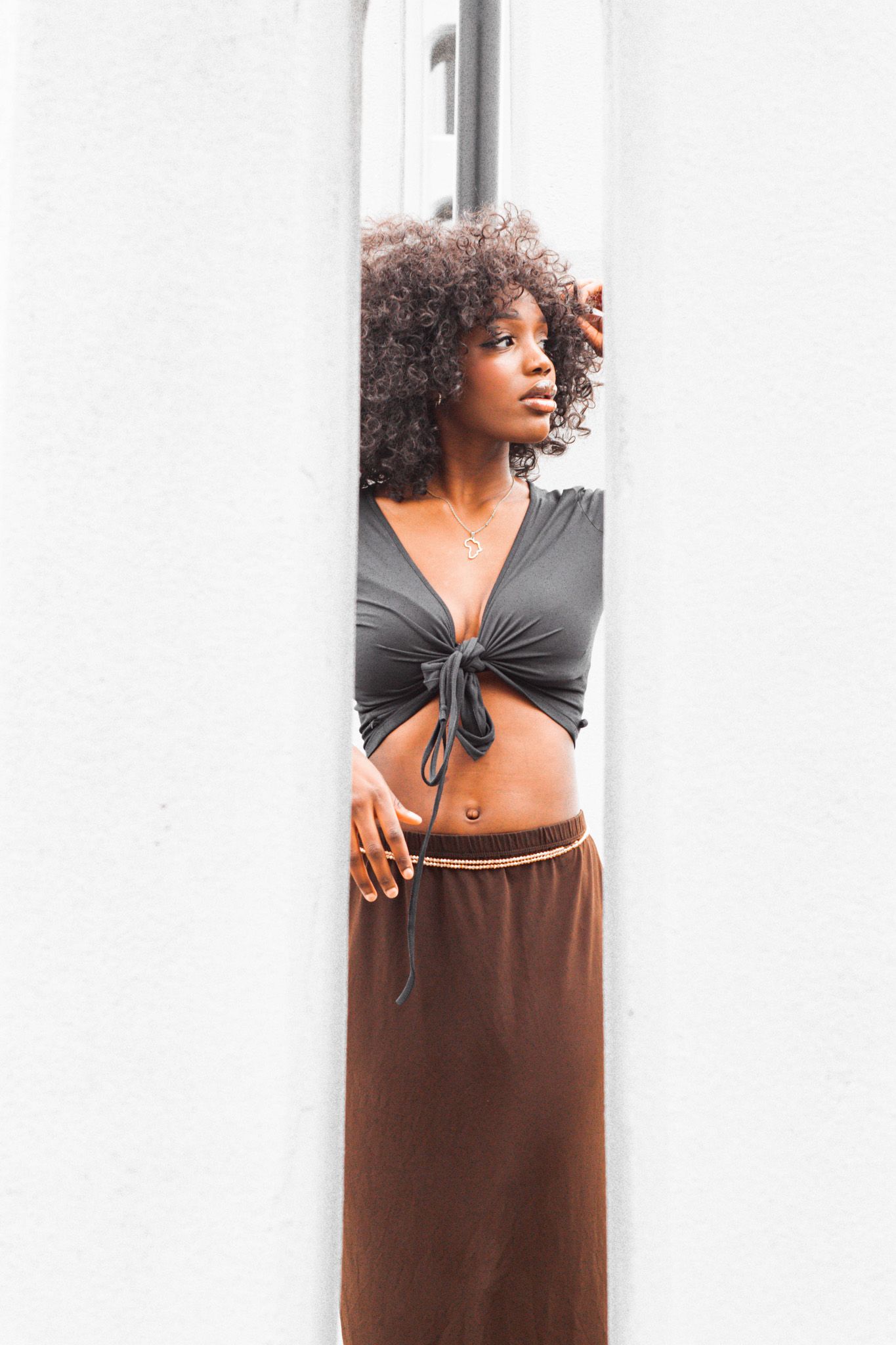 Model in gray crop top and brown maxi skirt poses against white wall with natural curly hair.