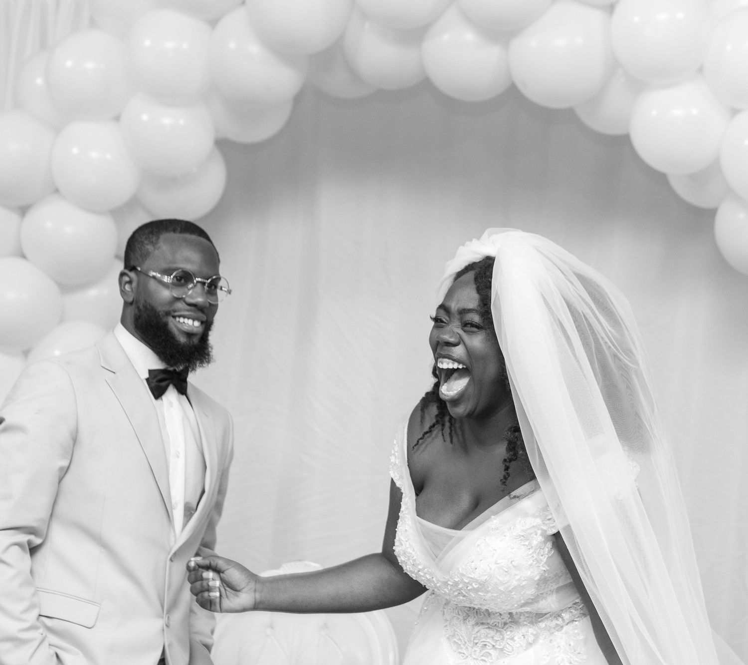 A joyful wedding couple shares a candid moment under a white balloon arch during their celebration.
